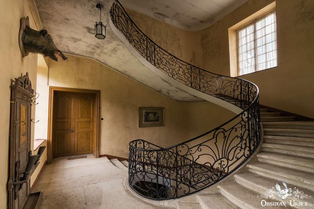 A grand, decaying staircase with ornate wrought iron railings in an abandoned castle, featuring a taxidermy boar head on the left wall.