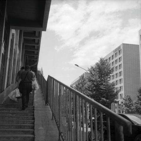 Lucky LUCKY SHD 100 (6x6)

English
A black and white photograph of a pathway that ascends to some overhead structure. Several people are climbing on the stairs, with things in their hands. The stairs have rails for protection. On the opposite side of the stairs across the street stands a group of buildings, halfway covered by some trees. The street seems vibrant with cars and pedestrians in sight. A streetlamp post is also visible slightly above the building top. The sky is bright yet cloudy.

…