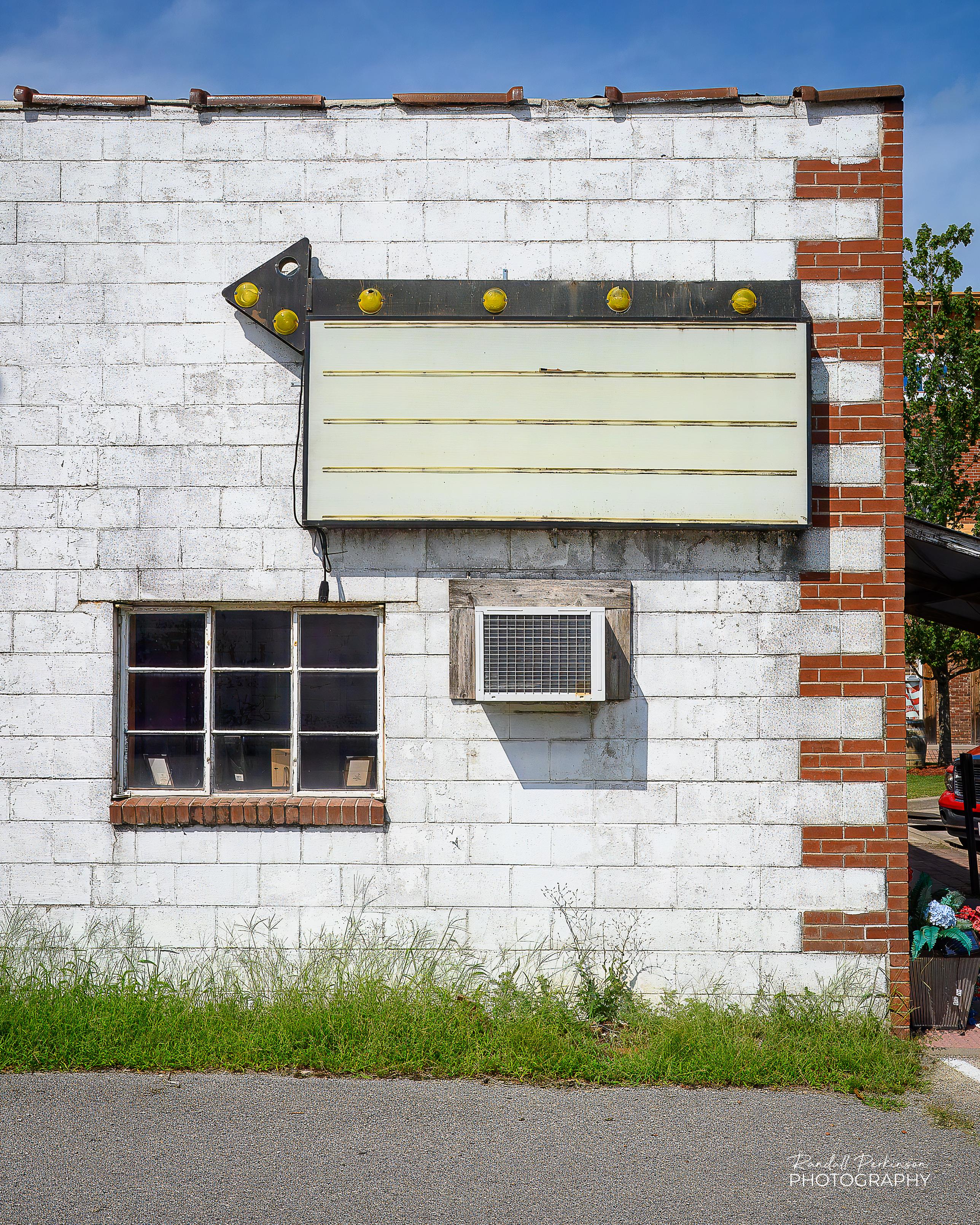 The side of a white concrete block building with red brick along the edge of the building.  There is one window, an air conditioner, and a sign with a black arrow and yellow lights and an area to place variable messages.