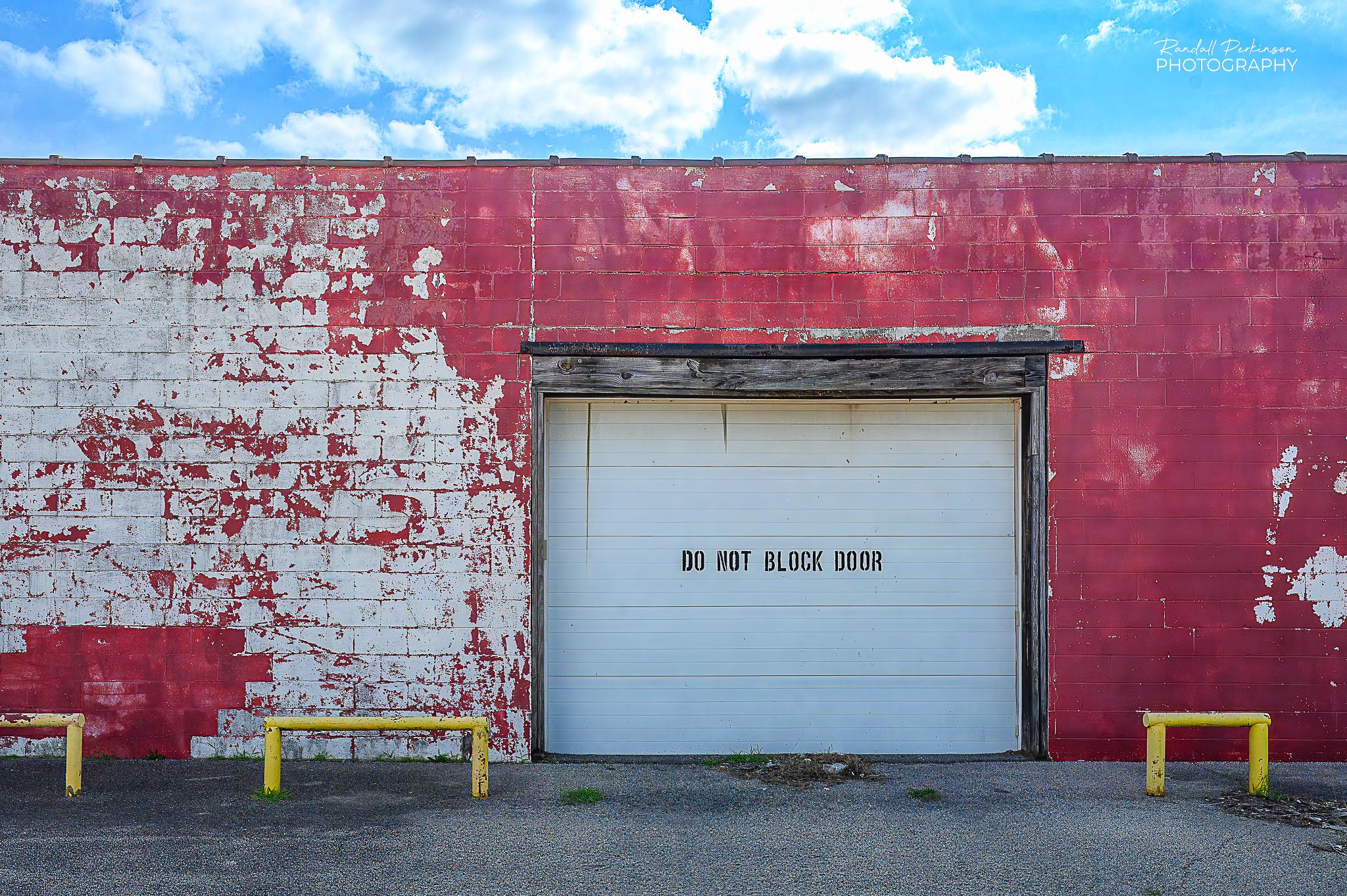 The side of a one-story concrete building with a garage door  with text containing the words "DO NOT BLOCK DOOR".  The red paint on the wall is peeling and weathering off to reveal white paint under it.  There are three yellow pipe bollards in front of the wall to prevent people from hitting the wall.