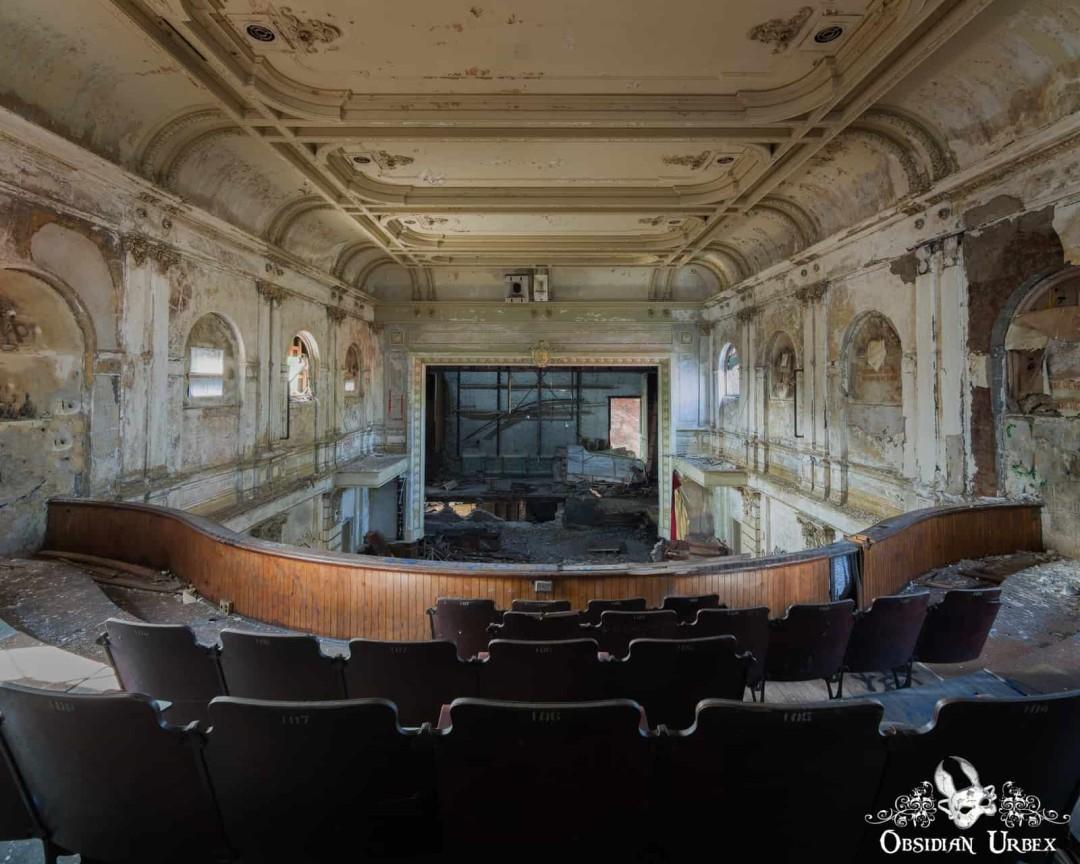 An abandoned auditorium, viewed from the balcony, with a collapsing stage and decaying walls
