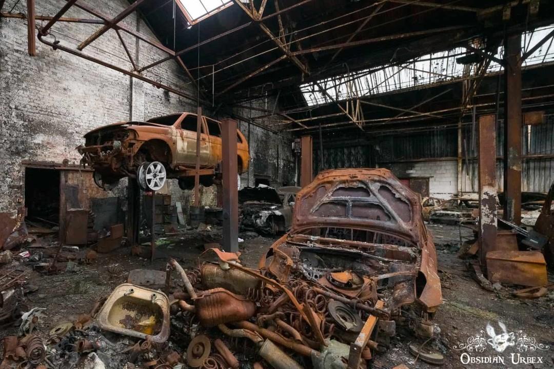 An abandoned, fire-damaged car dealership features a rusty car on a lift amidst charred vehicle wreckage.
