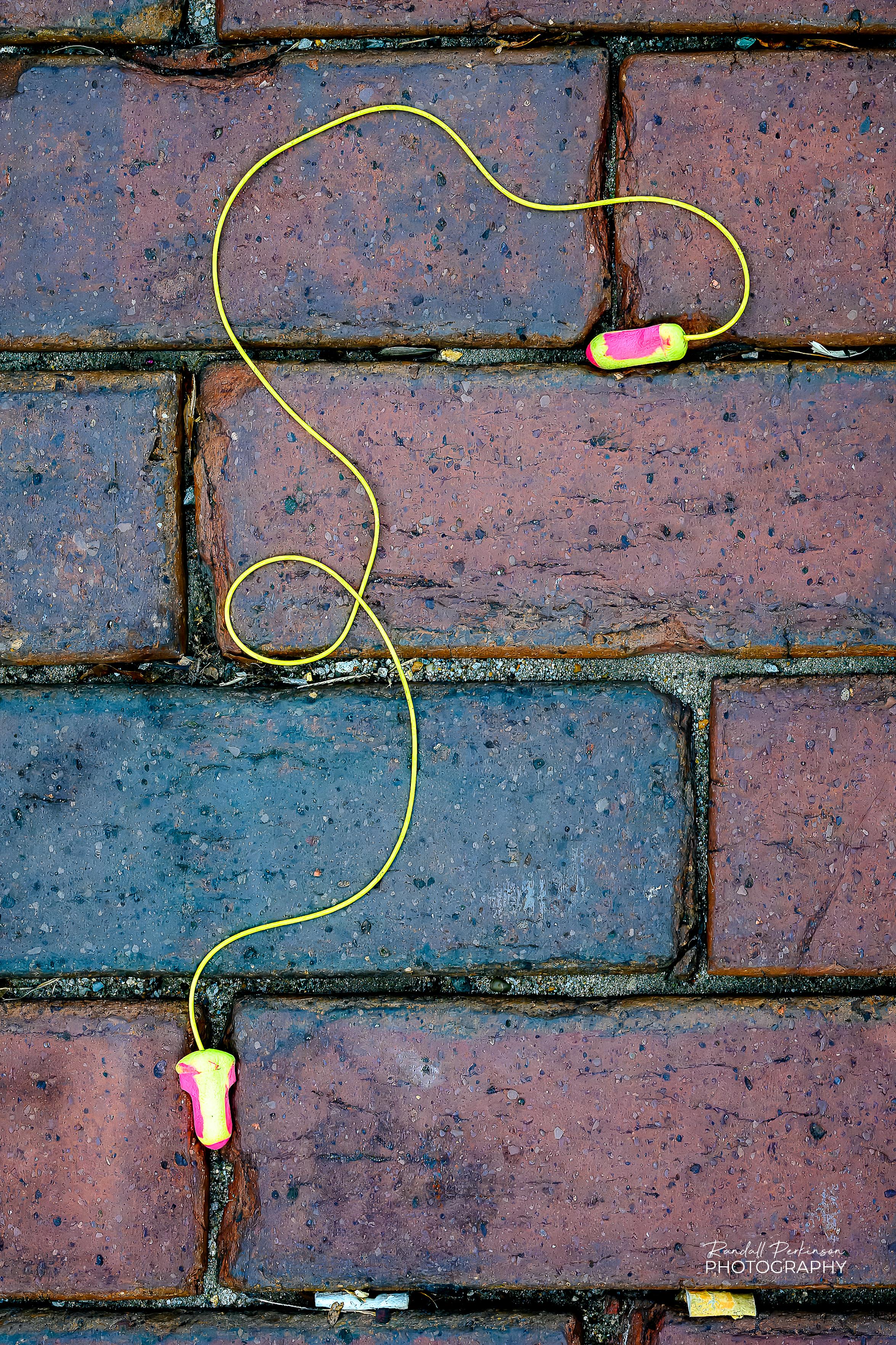 A pair of yellow and pink earplugs with a yellow cord connecting them laying on a brick street.
