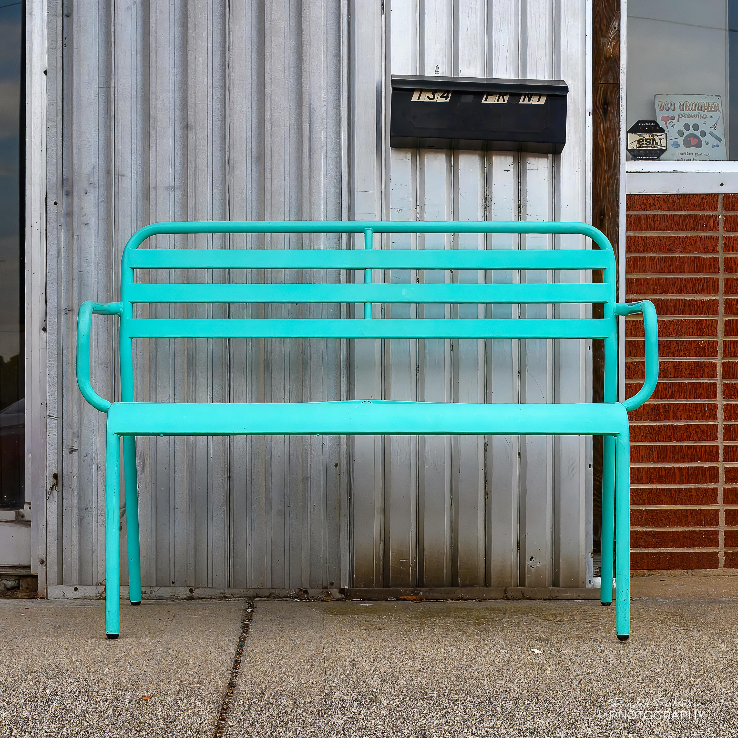 A lime green metal bench wide enough for two people to sit on it sits on a sidewalk in front of a building with silver metal siding and a black mailbox.