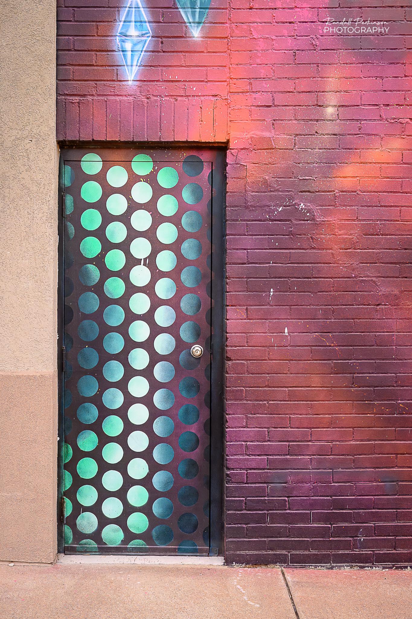 A metal entry door is covered in columns of dots of a gradient of green colors from very light to very dark.  The brick wall above and to the right are covered with a spray-painted design.