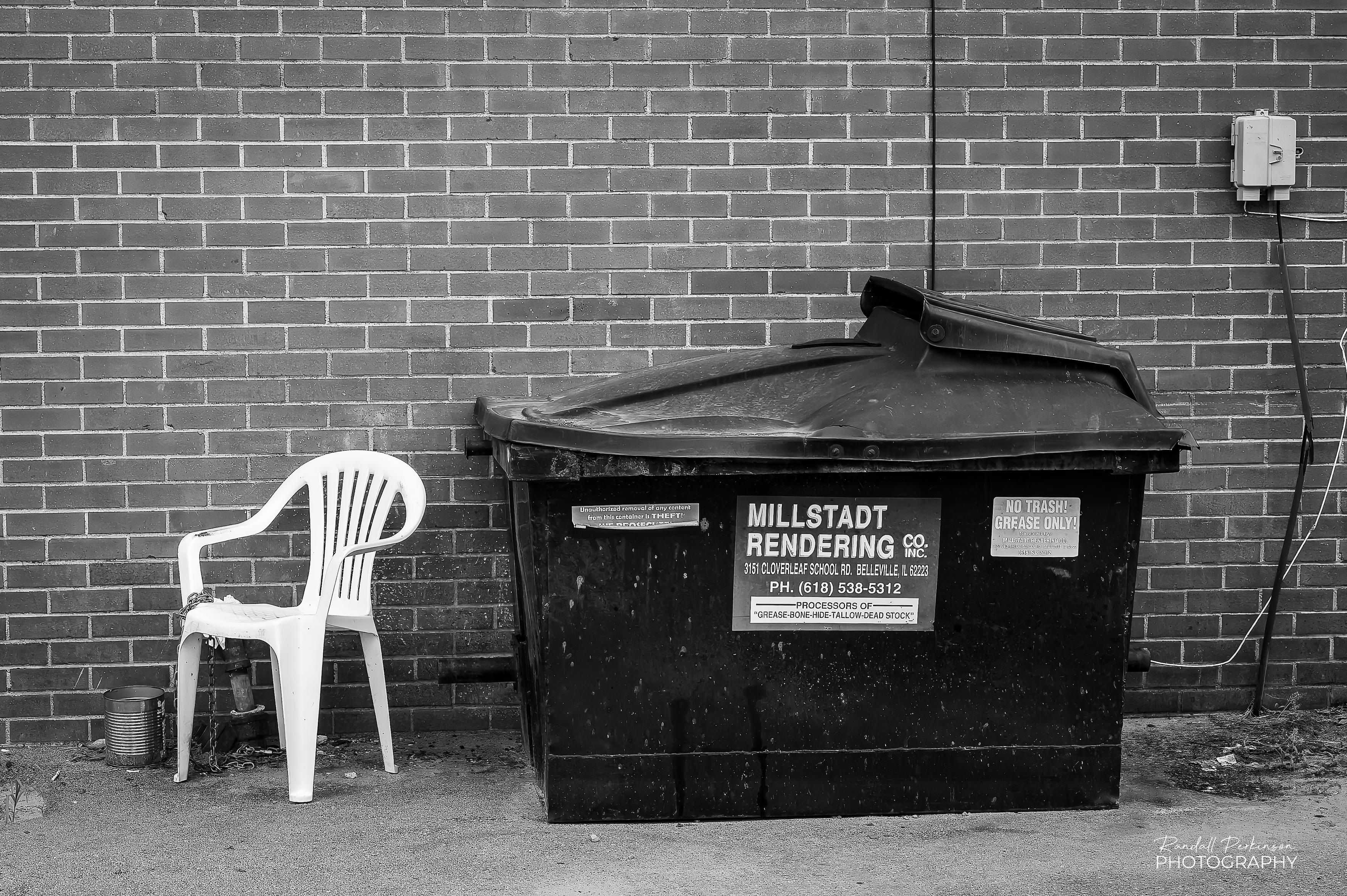A white plastic chair, chained to an adjacent pipe, sits in an alley next to a bin used for the collection of old grease.