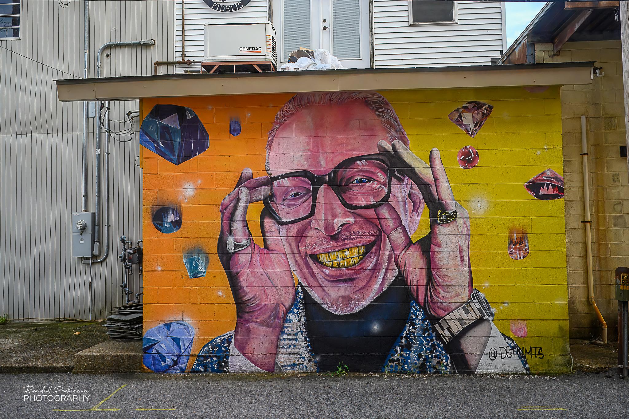 A large (about 8 foot tall by 12 foot wide) mural of the head and shoulders of a man wearing glasses with his hands up touching the glasses.  The background is orange and lime green and has diamonds and other gemstones floating around the man.