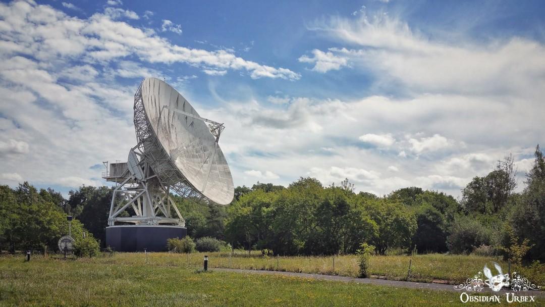 A large white satellite dish stands prominently in a grassy field under a bright, cloudy sky, surrounded by green trees.