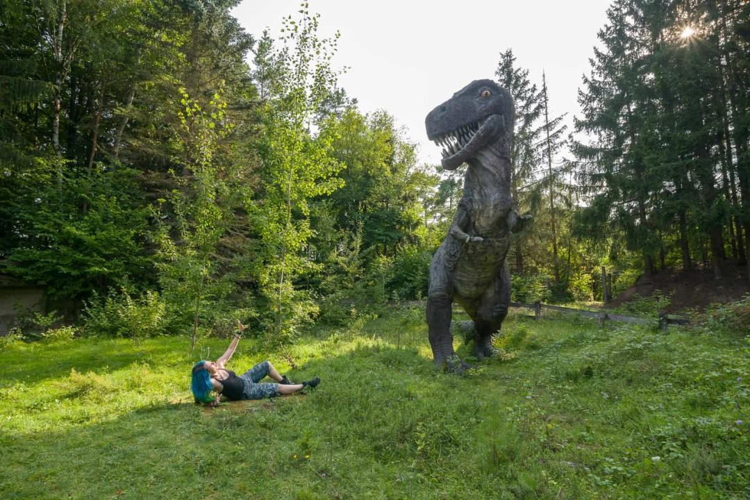 A person with blue hair lies on the grass, playfully interacting with a large T-Rex statue in a sunny outdoor park setting. The dinosaur stands tall with its mouth open, facing the person, surrounded by trees.