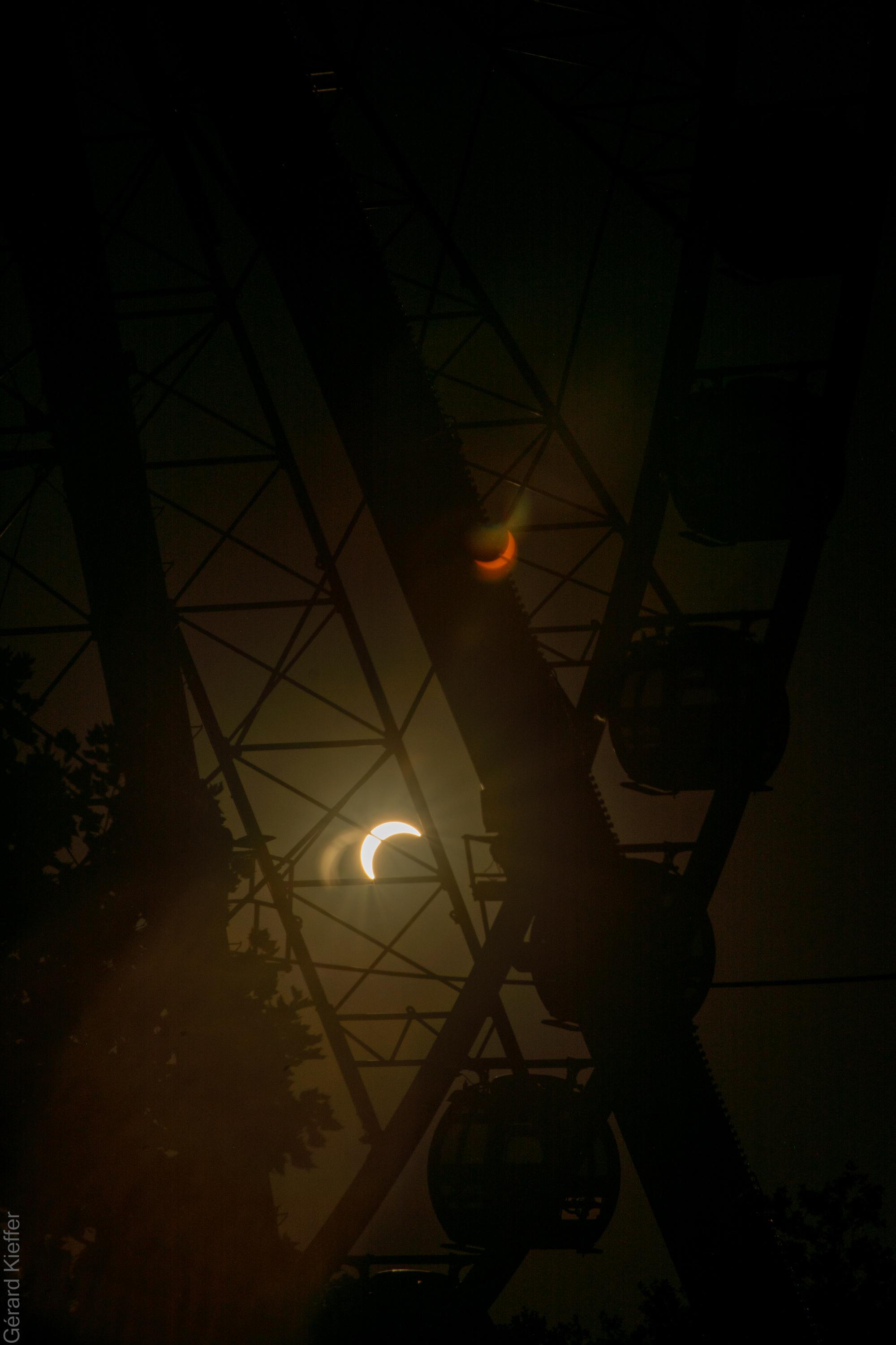 Solar eclipse seen through the framework of a Ferris wheel, with gondolas silhouetted against the dark sky. A lens flare creates a second, mirrored crescent eclipse in darker colors.