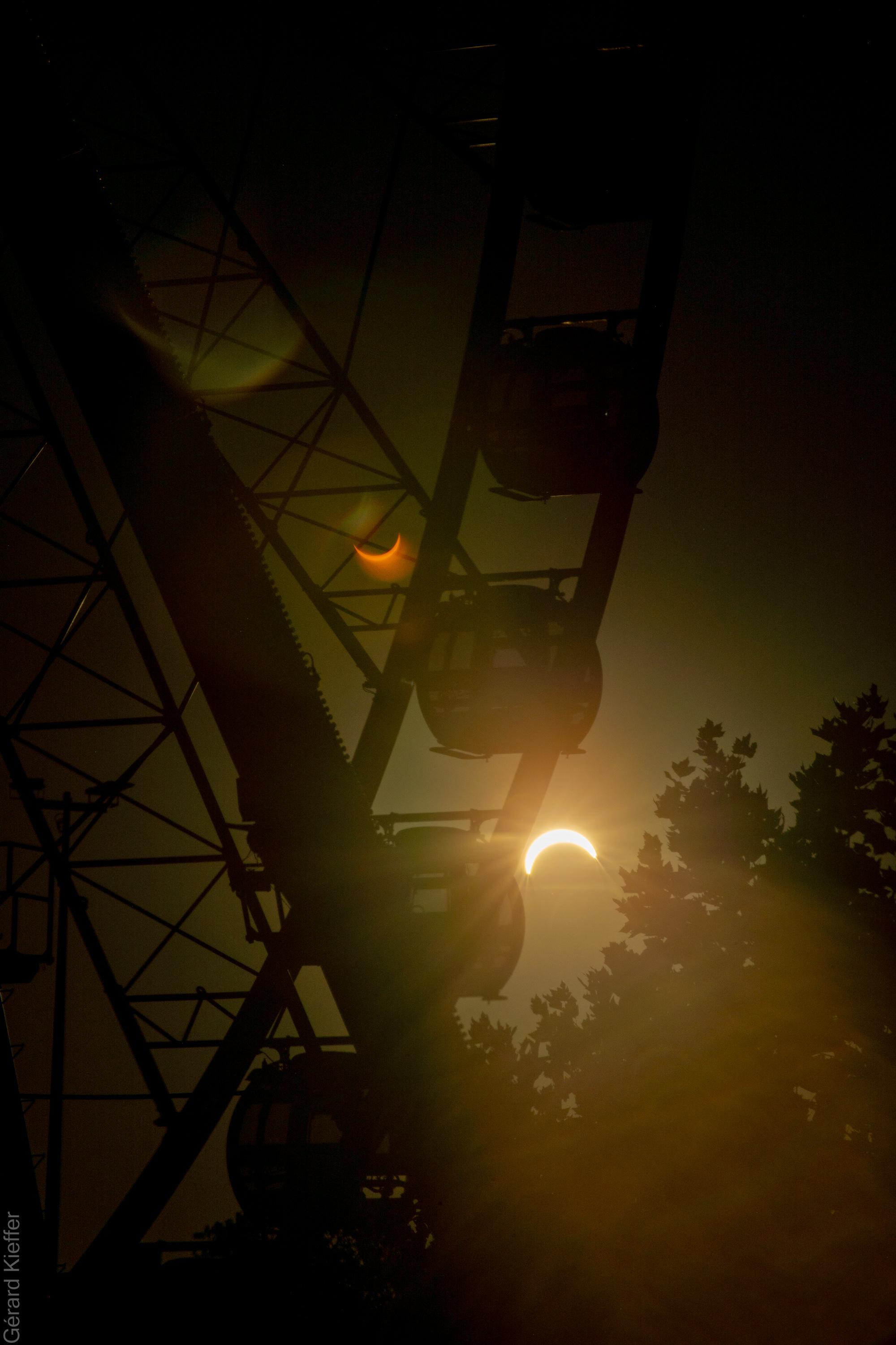 Solar eclipse seen through the framework of a Ferris wheel, with gondolas silhouetted against the dark sky. A lens flare creates a second, mirrored crescent eclipse in darker colors.