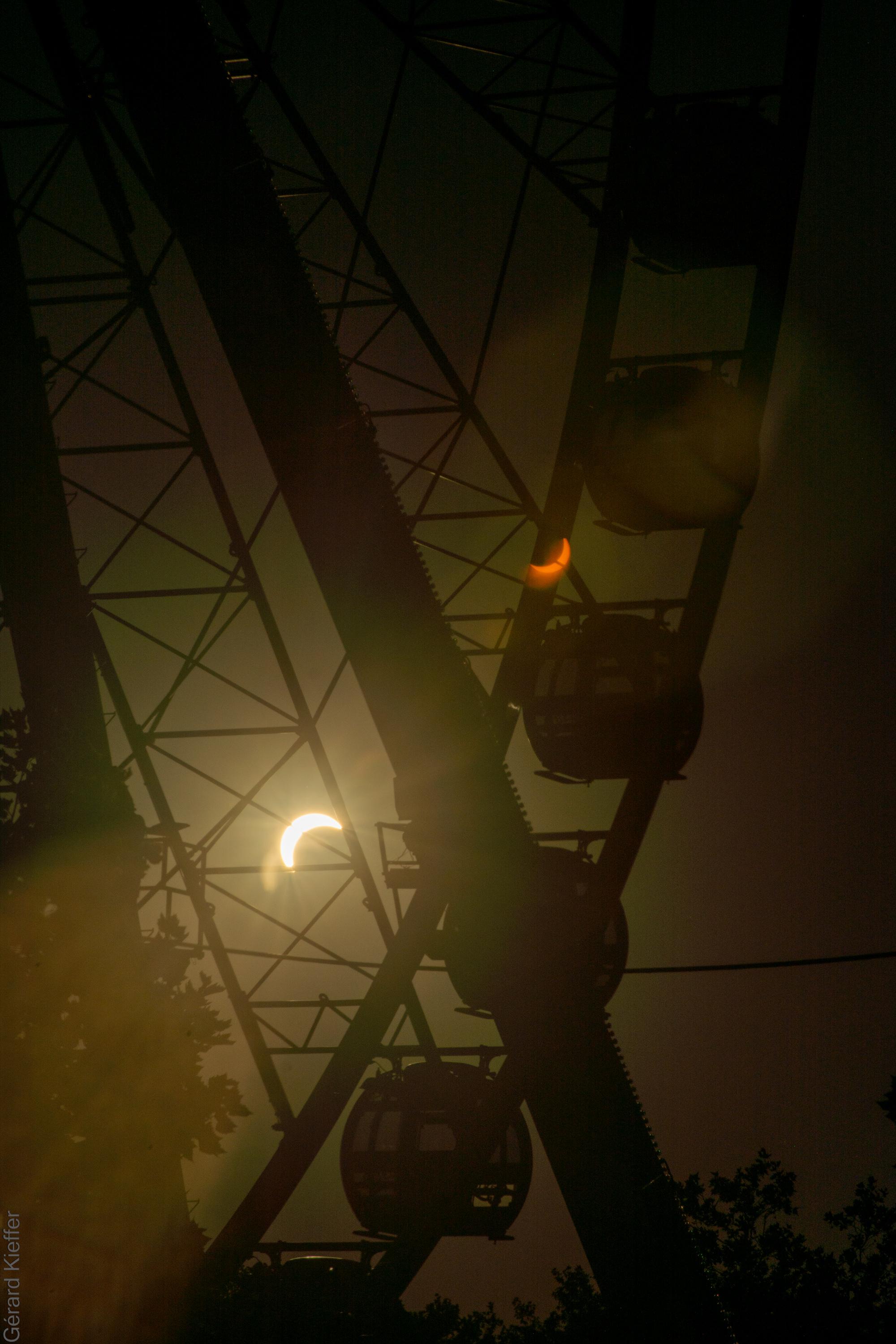 Solar eclipse seen through the framework of a Ferris wheel, with gondolas silhouetted against the dark sky. A lens flare creates a second, mirrored crescent eclipse in darker colors.