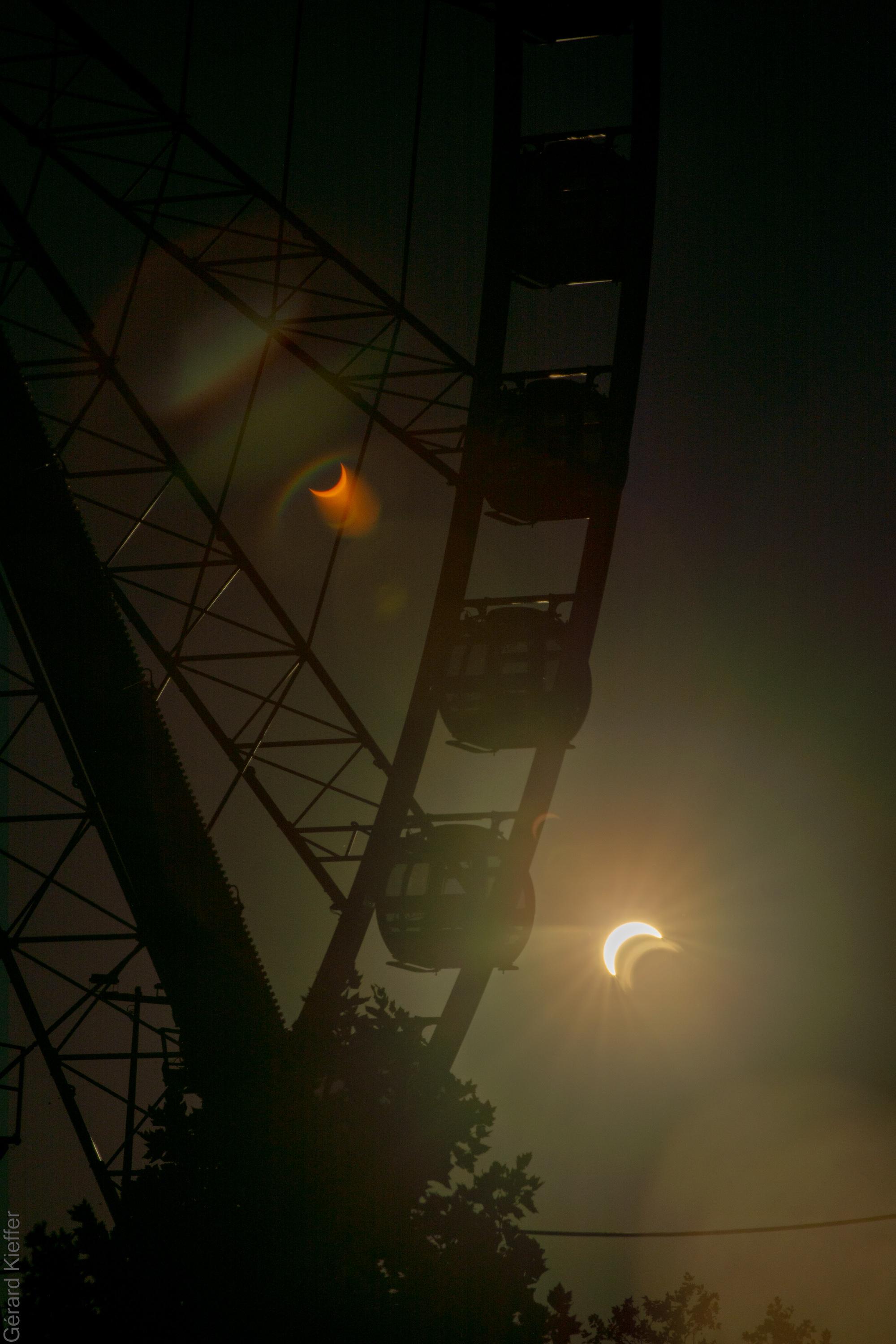 Solar eclipse seen next to the framework of a Ferris wheel, with gondolas silhouetted against the dark sky. A lens flare creates a second, mirrored crescent eclipse in darker colors.