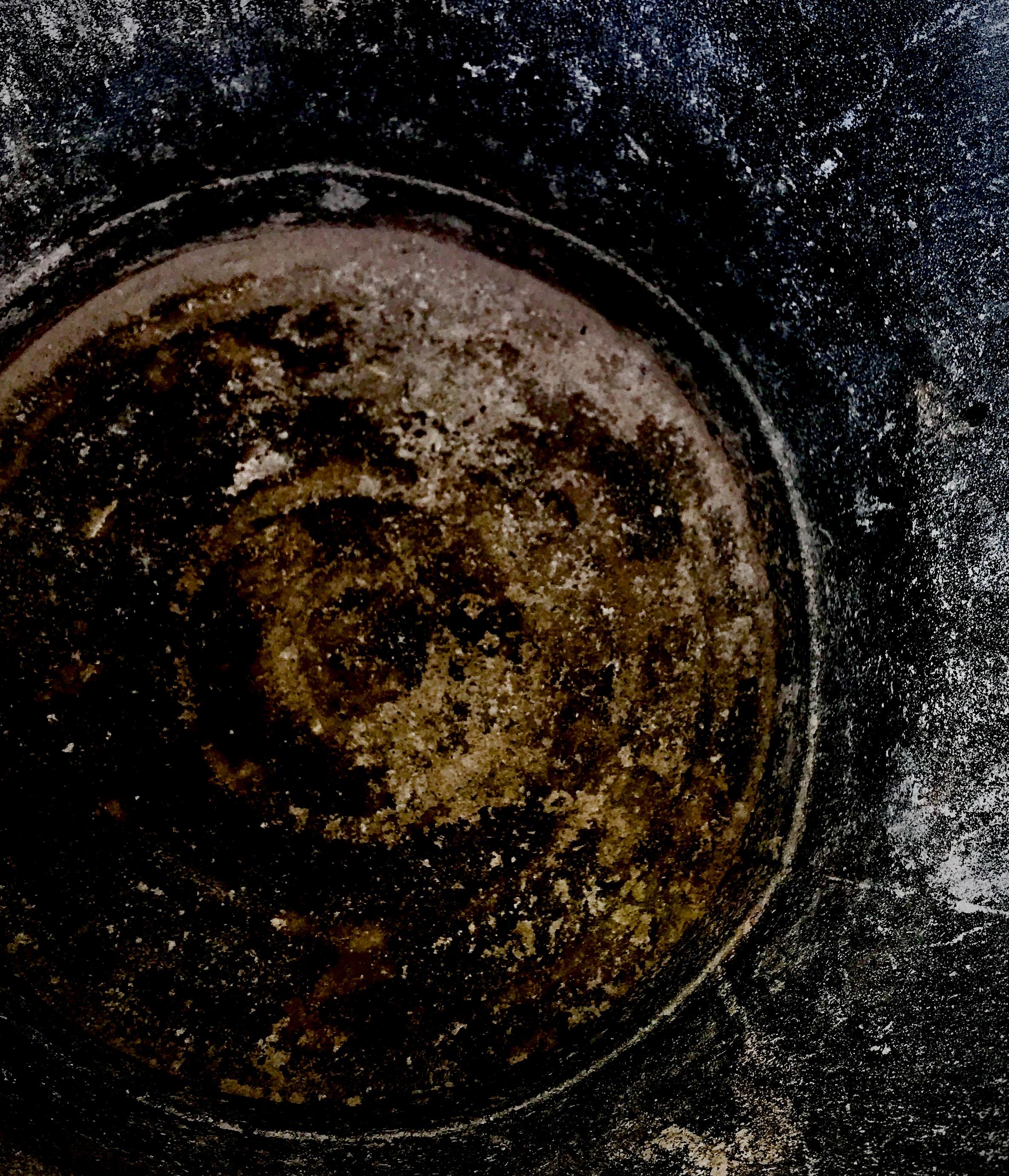Abstract, closely cropped photo looking directly at the bottom of a metal ash can. The sides and are dotted with carbon and light colored wood ash. The bottom has a dark umber hue, dotted with black carbon. I think it has the feel of outer space, filled with stars but containing a subtle geometry amidst the randomness.