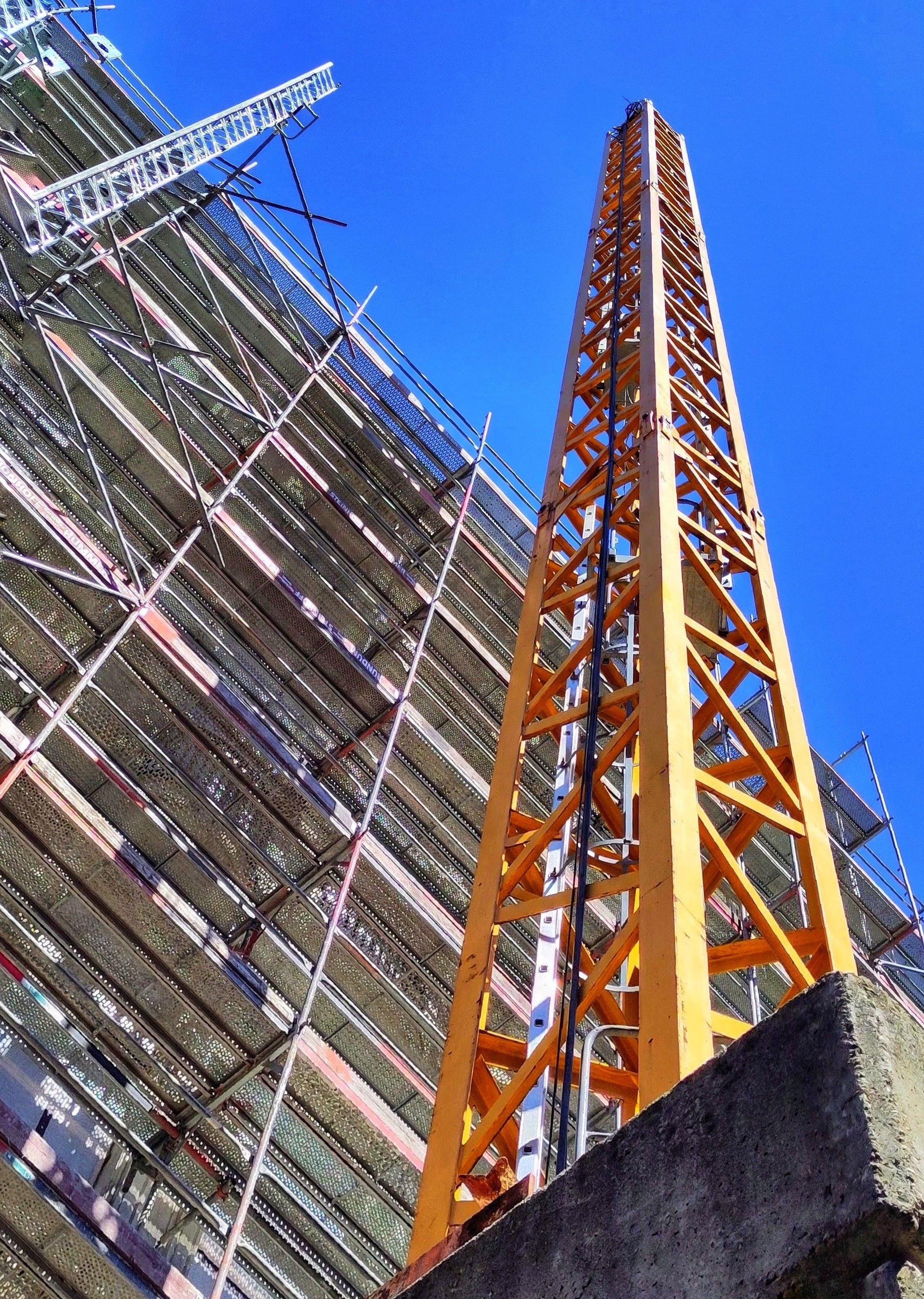The image depicts a construction site scene under a clear, vibrant blue sky. The perspective is from a low angle, looking upwards. Dominating the right side of the frame is a tall, bright yellow construction crane tower, viewed from its base, suggesting its immense height. The crane is constructed with a lattice of interconnected yellow beams forming a triangular, rigid structure. A dark cable or chain runs vertically along the inside of the crane's structure.
