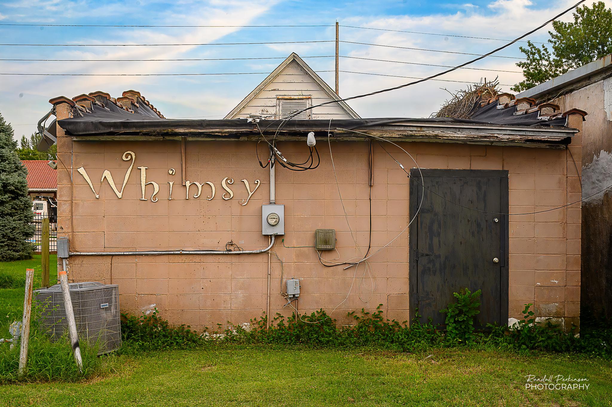 The back of a one story concrete block building painted a dull peach color with a black door.  On the wall of the building is a gold letters reading "Whimsy".