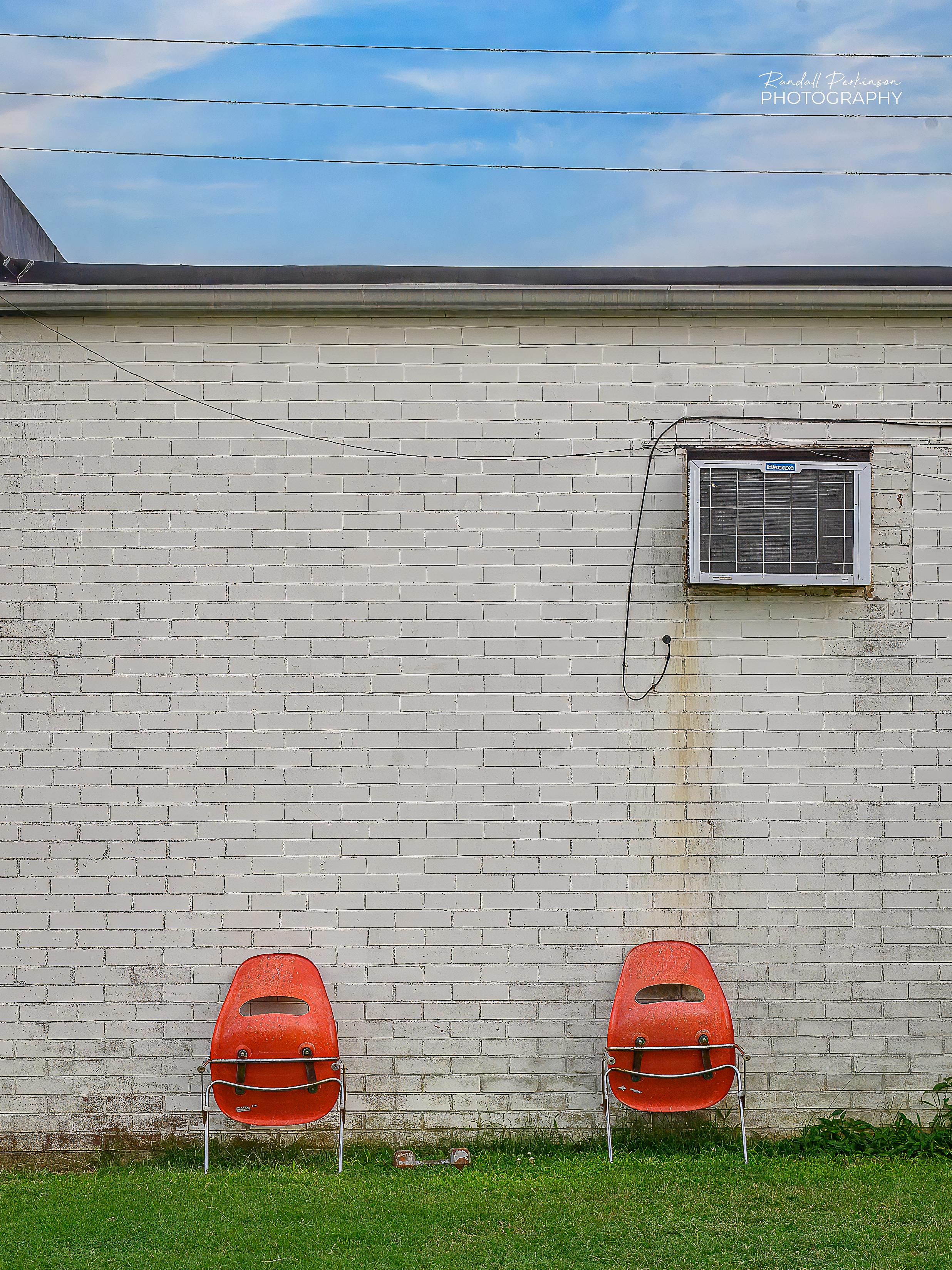 Two orange plastic chairs are leaned up against a brick wall painted tan.  There's an air conditioner mounted in the upper right portion of the wall.