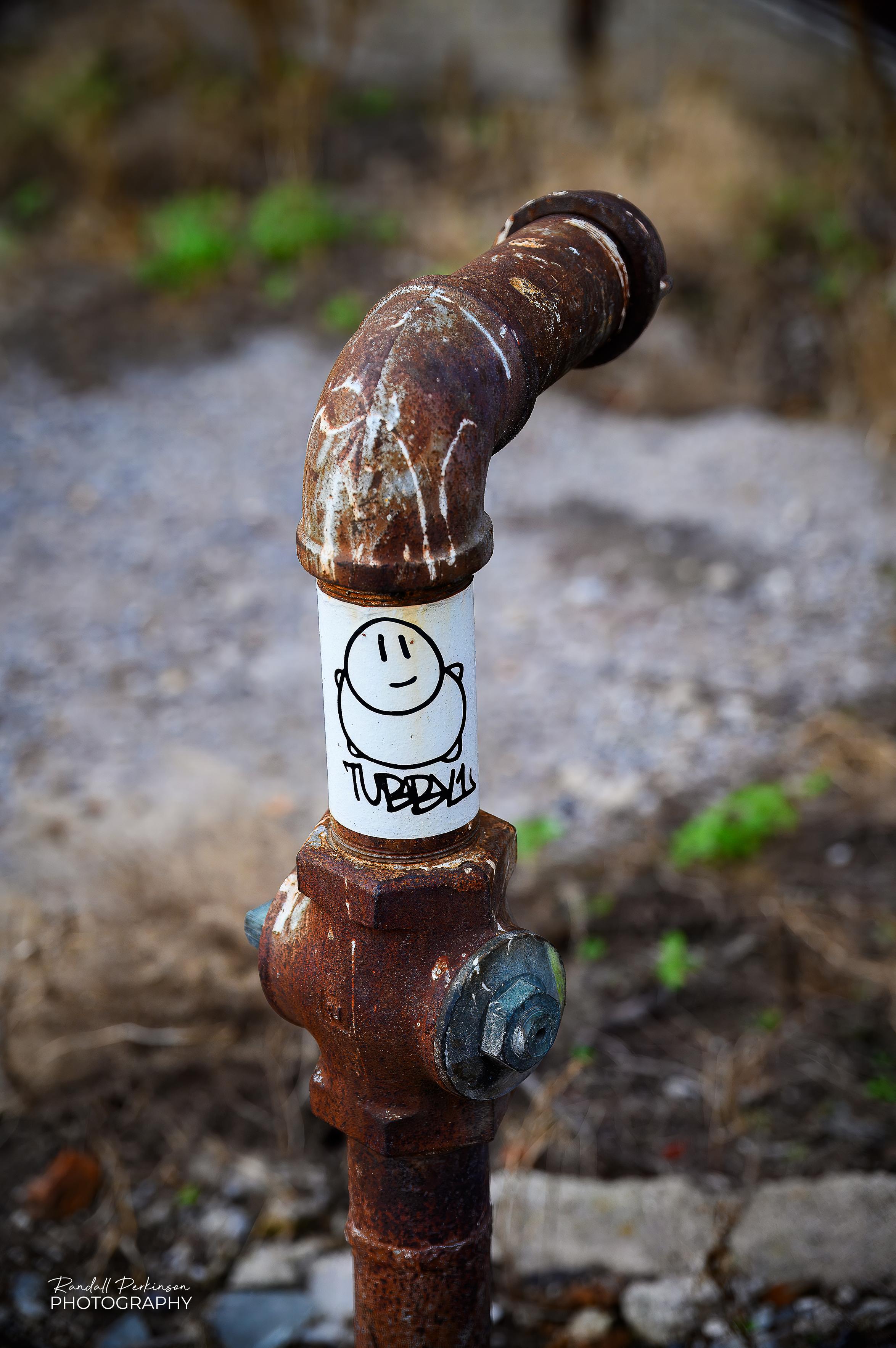 A large white sticker with a Tubby1 drawing and name is pasted around a rusty pipe sticking out of the ground in an alley.