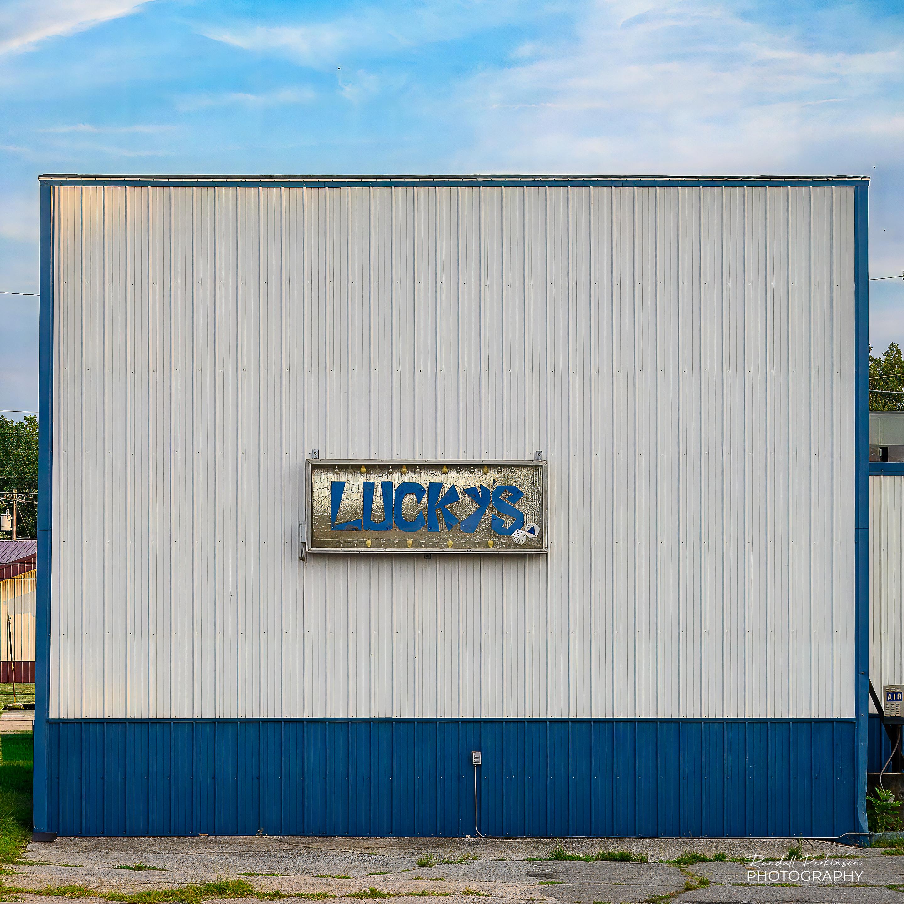 The side a building covered with white metal siding except for the bottom few feet that are covered in blue metal siding.  Not quite centered (really?) on the side of the building is a sign sying "LUCKY'S".