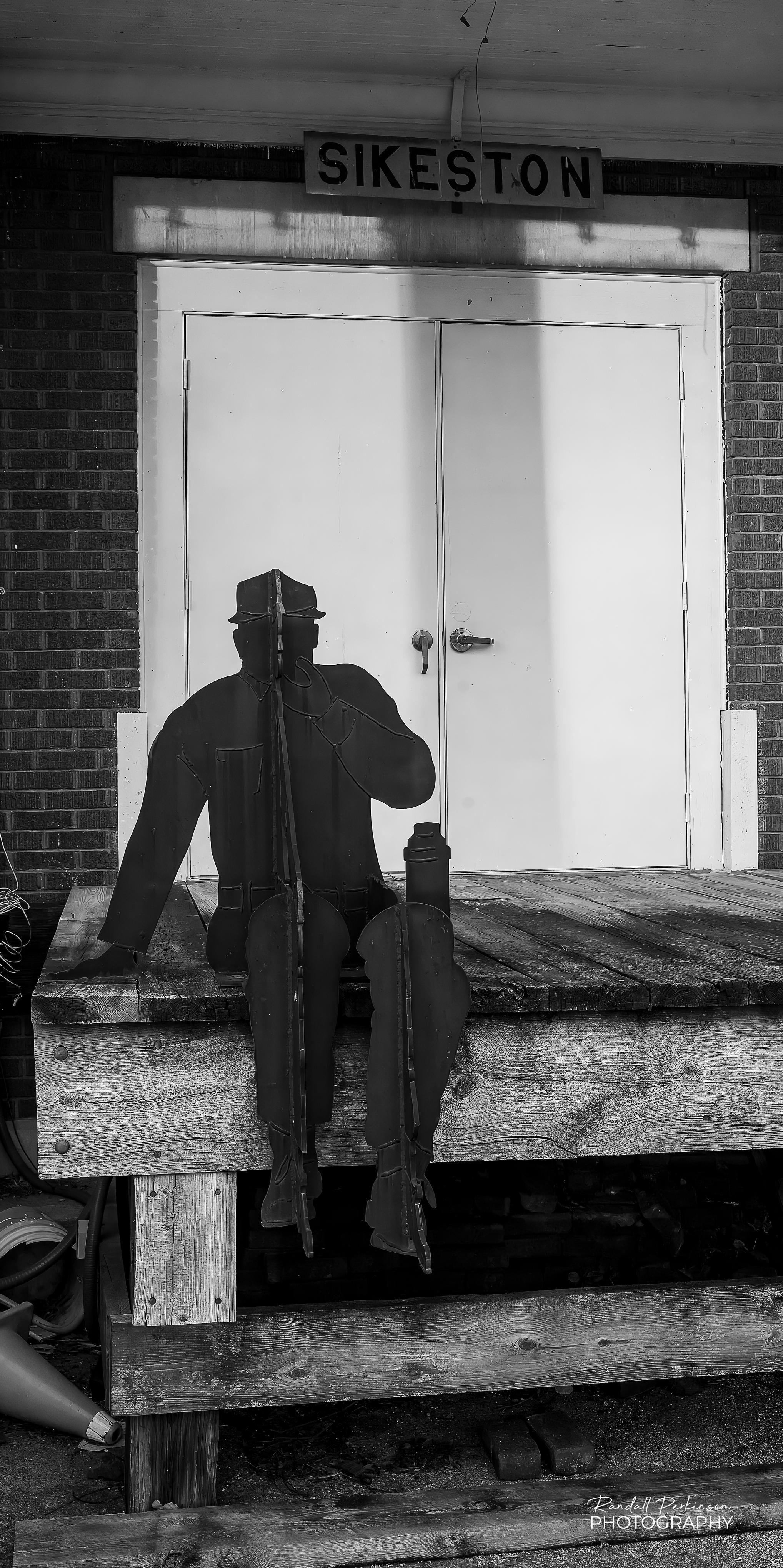 Multiple pieces of flat metal plates are cut in the silhouette shapes of people and mounted perpendicular to each other form the allusion of a railroad working taking a lunch or coffee break while sitting on a platform adjacent to an old railroad station.
