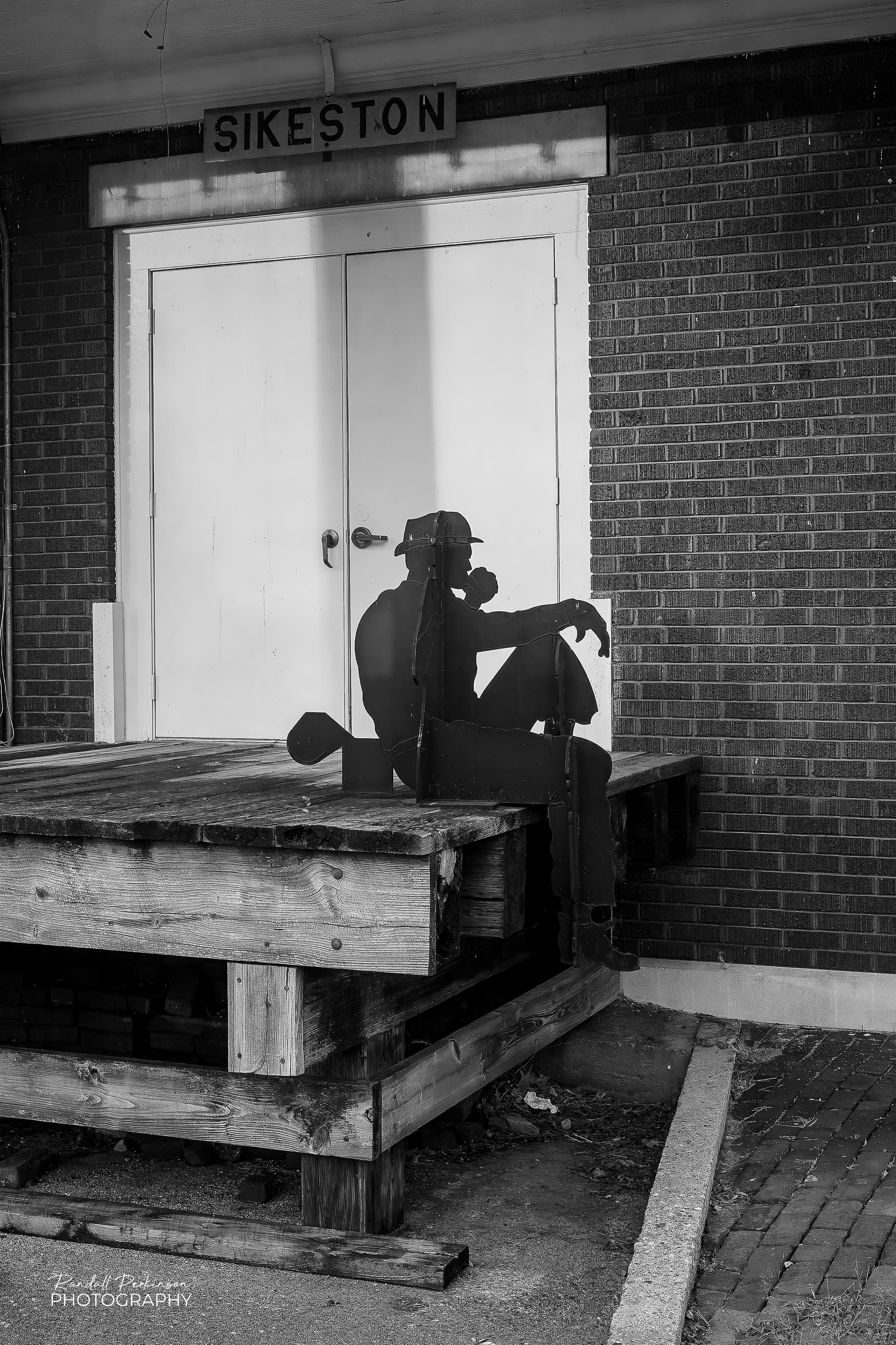 Multiple pieces of flat metal plates are cut in the silhouette shapes of people and mounted perpendicular to each other form the allusion of a railroad working taking a lunch or coffee break while sitting on a platform adjacent to an old railroad station.