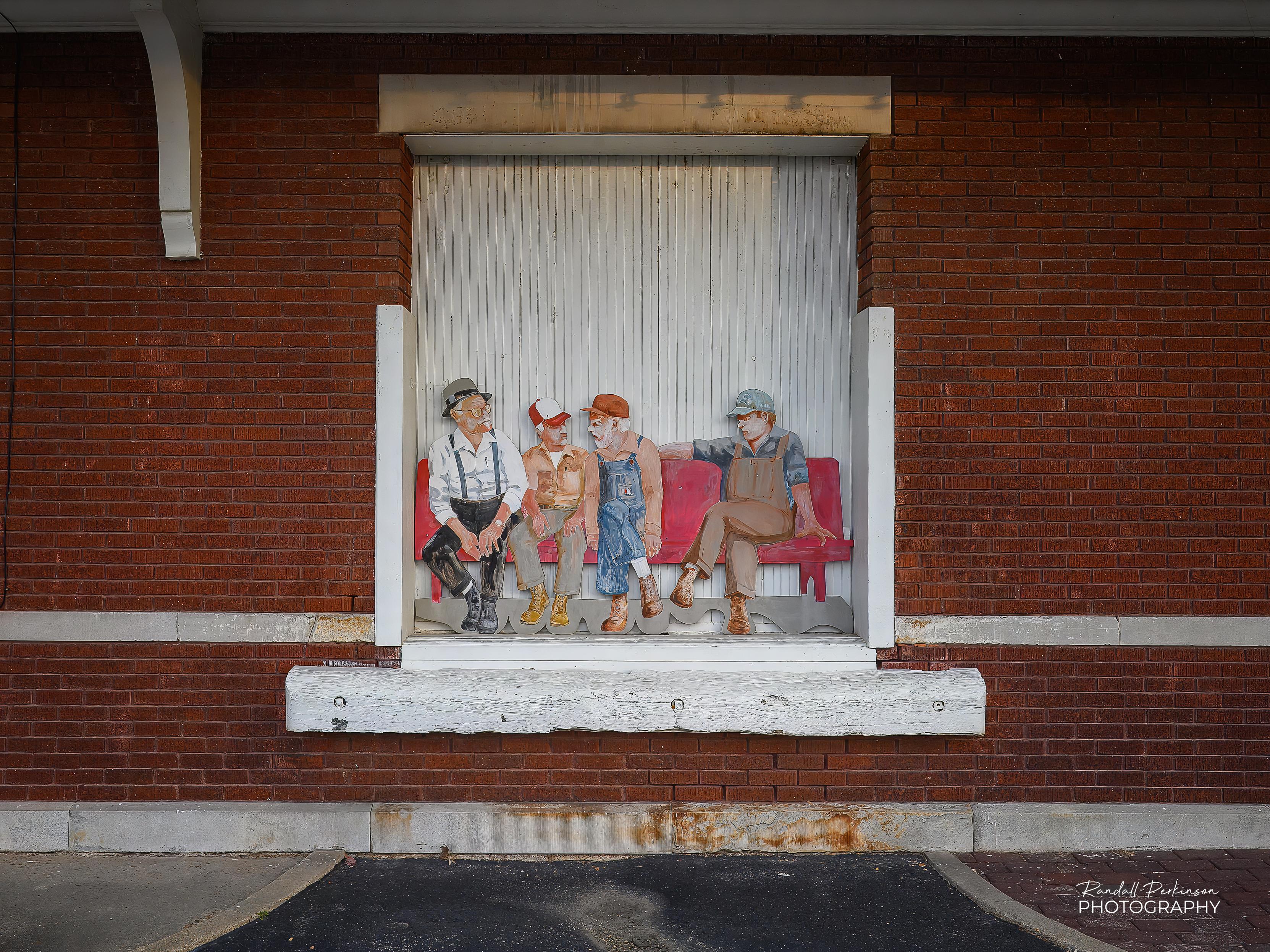 A view of a loading dock at an old brick railroad station with a piece of artwork showing 4 men sitting on a bench filling the width of the loading dock door.