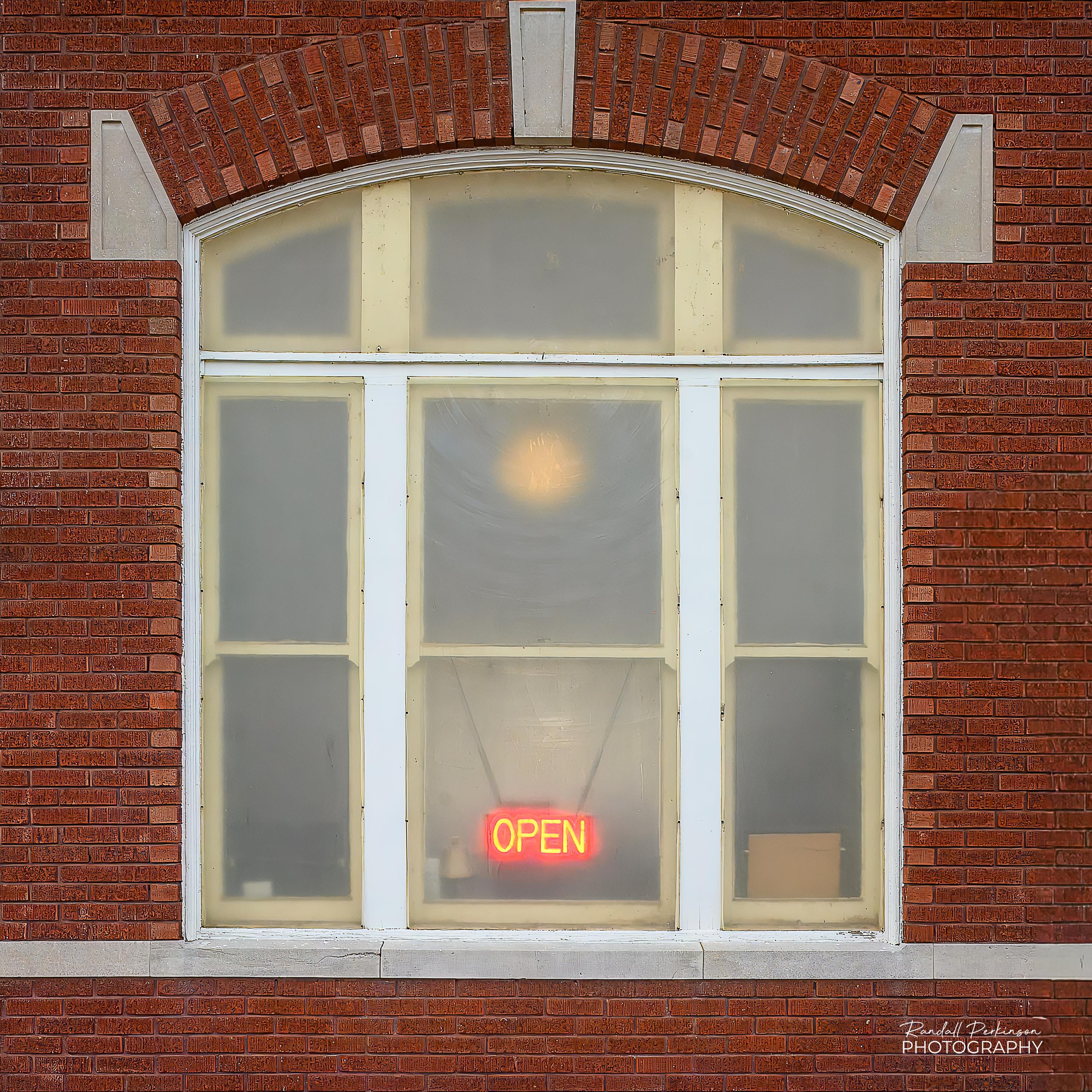 An arched window in an old brick train station with frosted glass.  Through the glass you can see a neon sign that says "OPEN" hanging in the center bottom window.