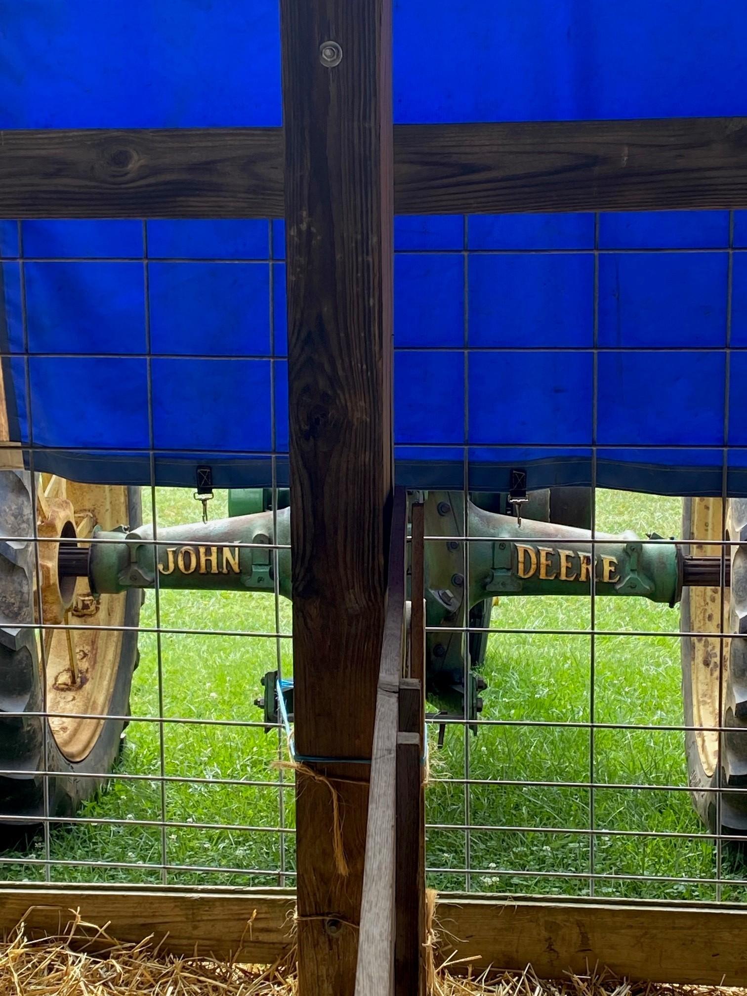 Color image of a vintage John Deere tractor parked outside a show barn at the Medina County Fair, Ohio, USA. Image shows the rear axle of an early John Deere farm tractor with the words "JOHN" and "DEERE" in yellow block type on both sides of the green axle housing, the large rear tires over yellow rims visible left and right, bright green summer grass below.