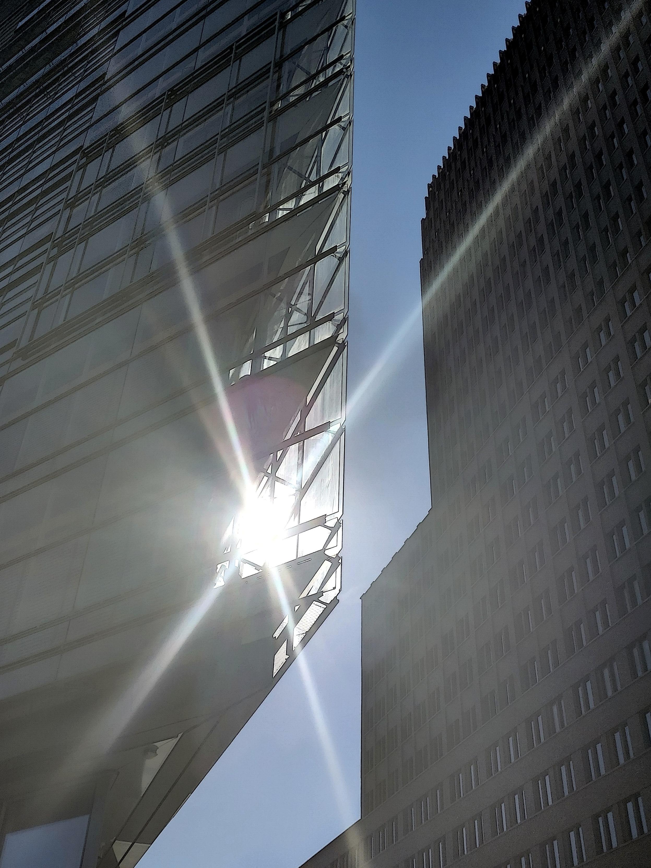 An upward view between two tall buildings against a clear blue sky. On the left is a glass-and-steel facade with an angular, irregular framework of struts; on the right, a grid of small windows on a lighter, flatter facade. The sun shines through the left building's structure near the centre, creating a bright starburst flare with long diffuse rays streaking diagonally across the frame and a soft lens-flare spot. The overall image is high-key and slightly hazy from the backlight.