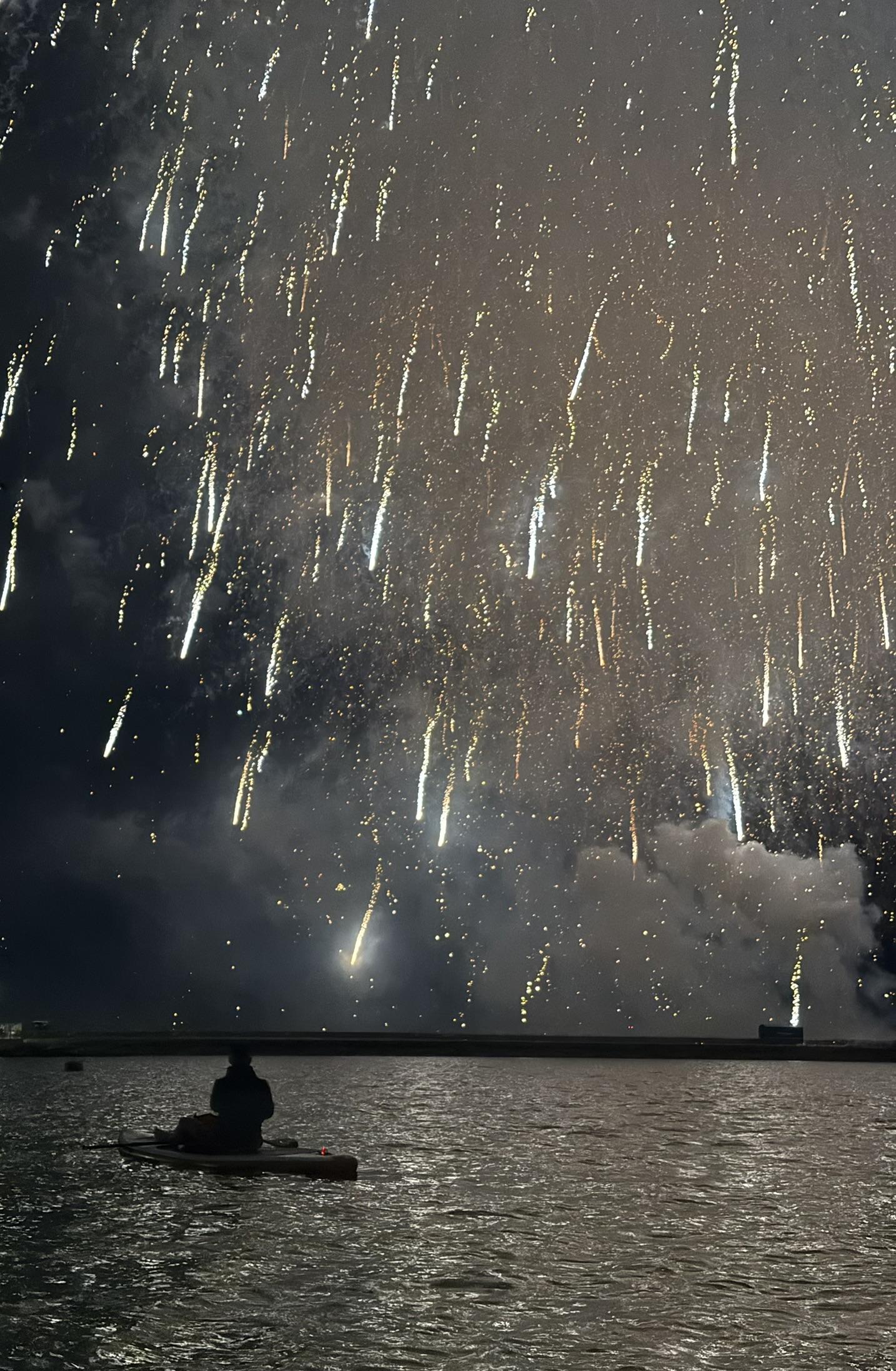 A firework in the sky, with a silhouette of a man sat on a paddleboard in front