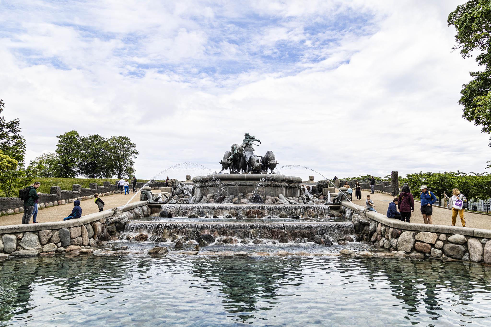 It's a bright day with lots of thin, white layers of cloud in the sky but plenty of patches of blue tearing them apart. We're looking at a fountain set back in a large pond either side of which there are wide paths that run along then rise up behind the fountain a few metres giving visitors good opportunities to see the structure from all around and at slightly different elevations. The pond has walls of large, rounded stones of varying sizes and rounded stones also punctuate the water's surface at the base of the two small, but wide cascades that step up to the fountain itself. The fountain is mounted on a curved, stone base practically as wide as the pond itself and the sculpted forms on the top are of a woman driving oxen directly towards us. Either side of the upper cascade there are thick serpents. The lower cascade is flanked by men's torsos, gripping the walls with their arms and turning their heads back to shoot jets of water over the pond, crossing over just before they hit th