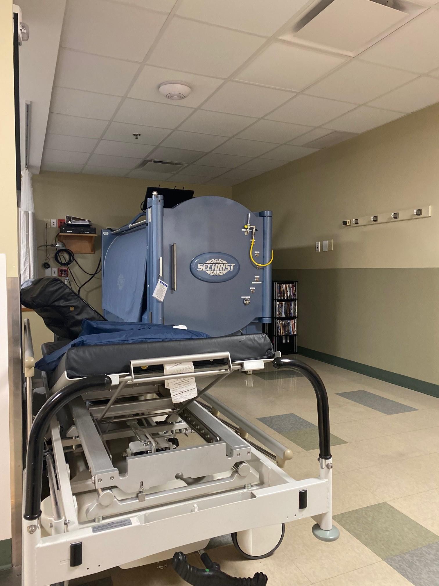 Color image of a Sechrist hyperbaric chamber. Image shows a draped hyperbaric chamber located in a hospital unit treatment room, with the feeding gurney next to it, nearer in the image; the gurney feeds the patient into the opened door of the chamber, which is then closed and sealed, with the atmospheric pressure then modulated by the operator in a 100% oxygen environment to treat the patient.