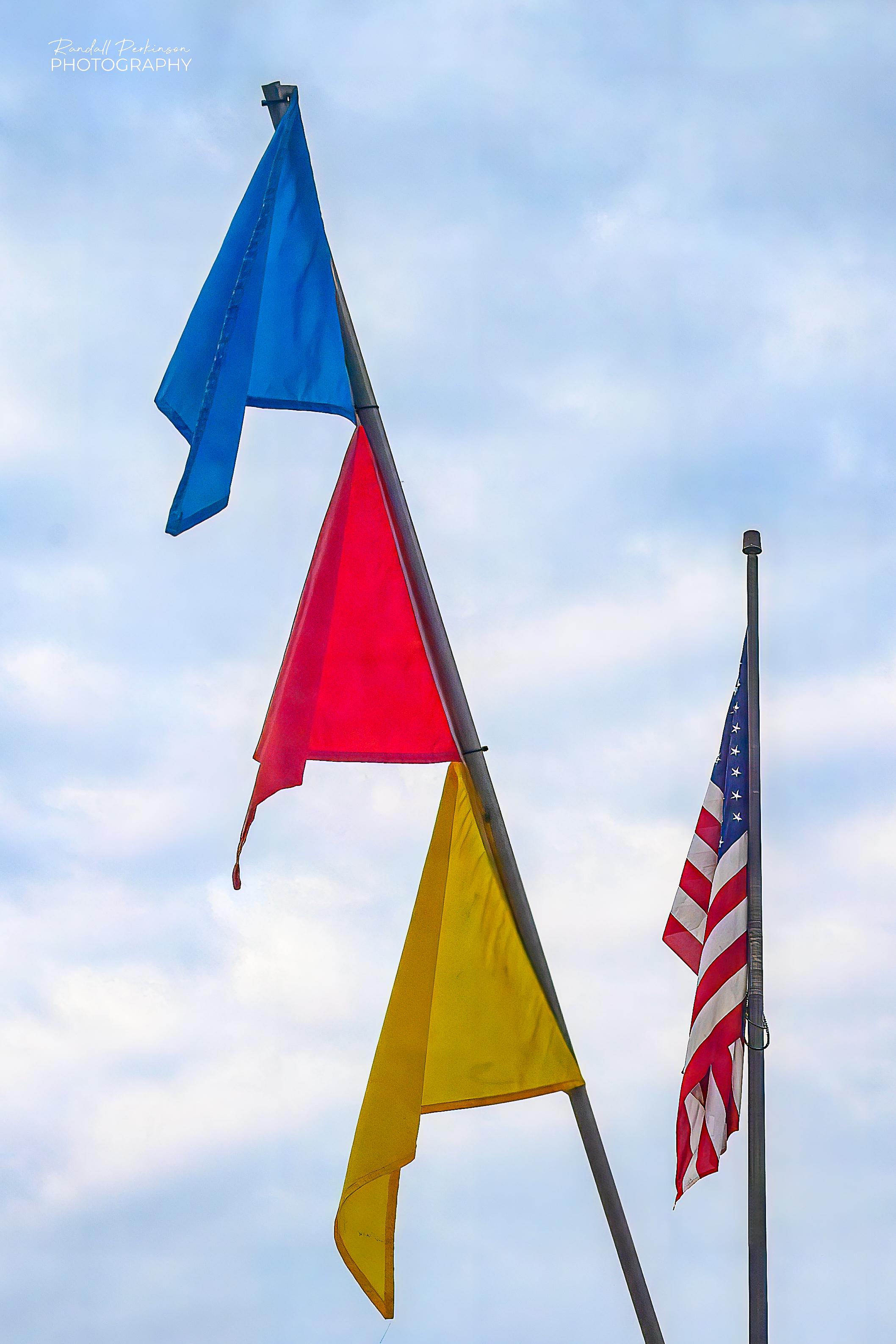 A view of a blue, red, and yellow flag on one pole and in the background the US flag on a flagpole.  Nothing but cloudy skies behind them.