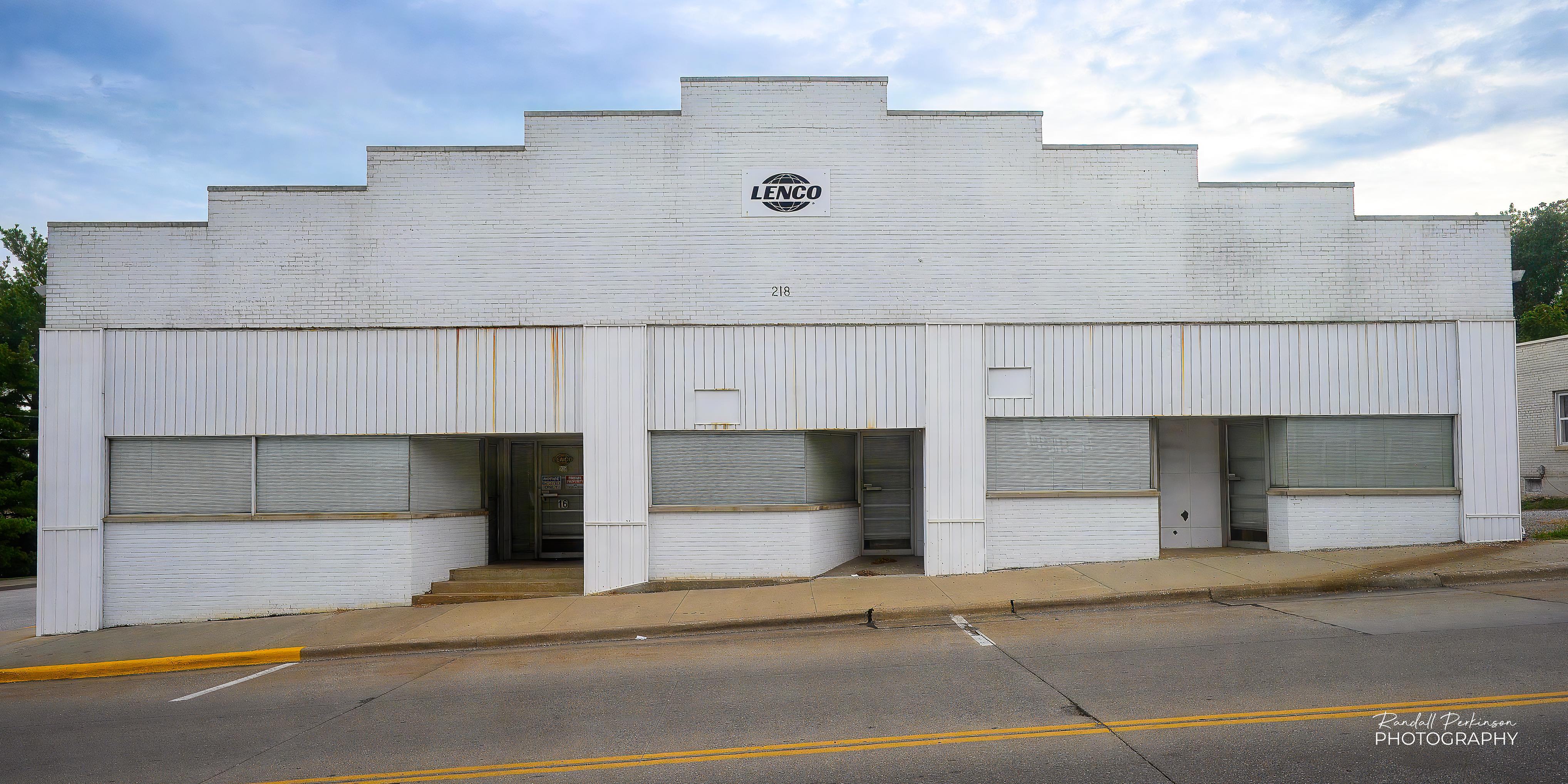 Front view of a one story brick building painted white.  The roofline steps from each side to a high point in the middle.  There are three separate entrances.  The building sits beside a street that is on a hill rising from left to right.