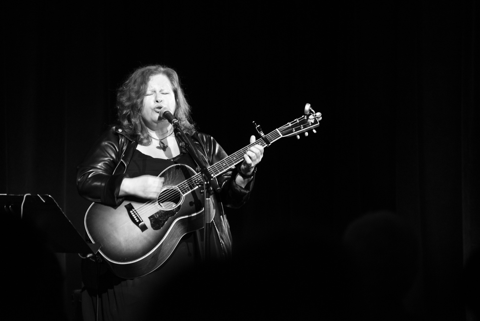 A black and white shot of Suzie playing acoustic guitar and singing on stage at Hugh’s Room Live. The acoustic bears the scars of years on the road.