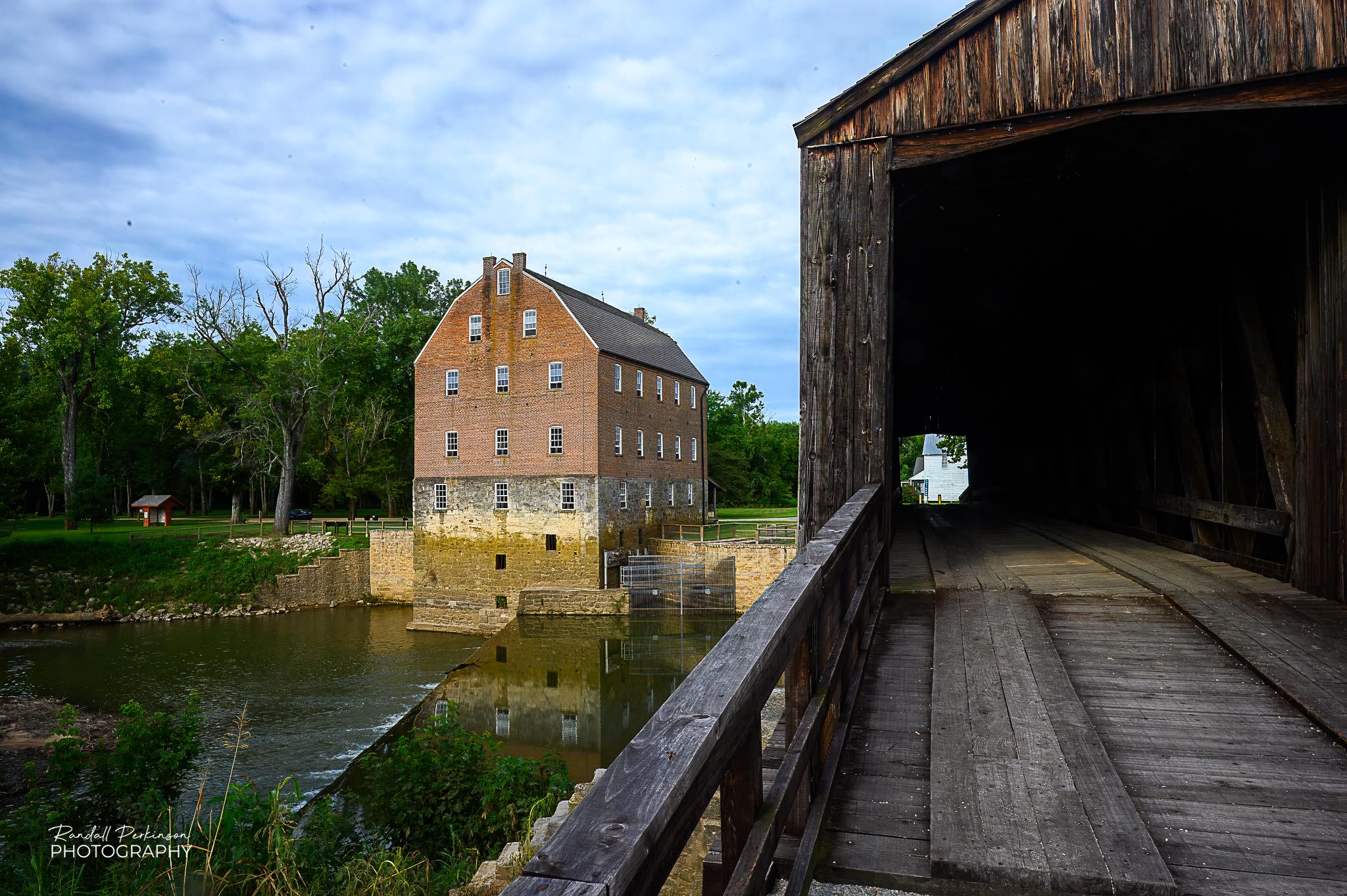 The right half shows a view down the length of a covered bridge.  The left half show an old stone and brick mill along the bank of a river.
