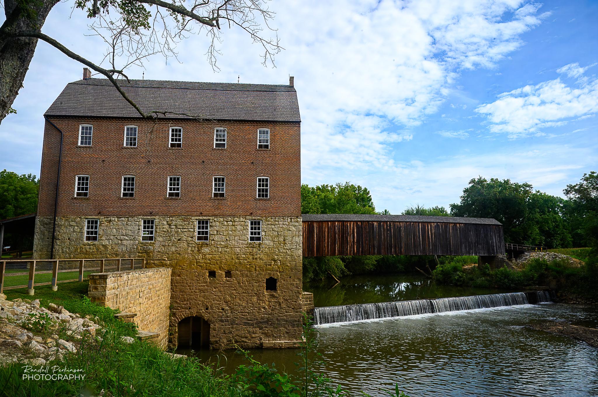A view of a multi-story stone and brick mill building and behind it a covered bridge.