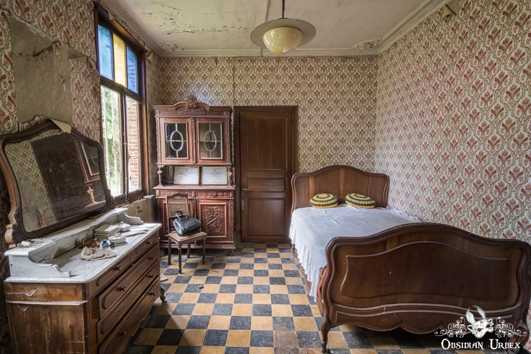 An abandoned bedroom with vintage wooden furniture, patterned wallpaper, and a checkered floor. A dresser with a mirror, bed, and cabinet are visible.
