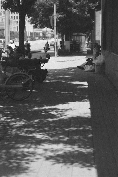 Ilford HP5 Plus 400 (FF)

English
A black and white photograph of a sidewalk. Two people can be seen resting on the side walk in the shade. Shadows of trees are cast on the sidewalk and some bicycles and motorcycles can also be seen. In the background, a bustling urban scene is seen. 

中文
一张人行道的黑白照片。两个人坐在阴影中休息。树荫在人行道上形成阴影。还能看到自行车以及摩托车。远处是城市的景观。