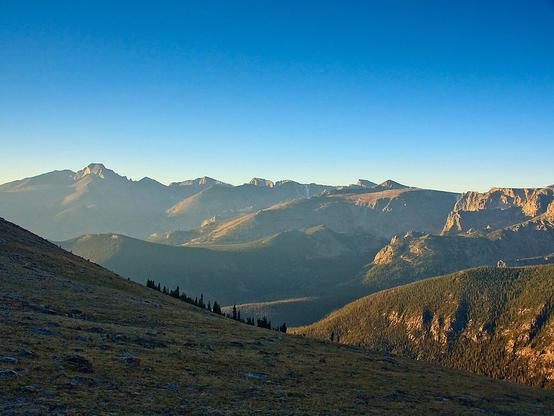 Panoramic photo in Rocky Mountain National Park. Hiking along the Ute Trail, looking East thru the gap at Longs Peak at the left and the rest of the Front Range extending to the right. Sunrise is just a few minutes ago but in November that’s not so early. Clear blue sky above with pale haze over the plains to the East. So quiet, at around 10-11,000 ft, 3000-3500m, and no wind, fortunately. November 2013.