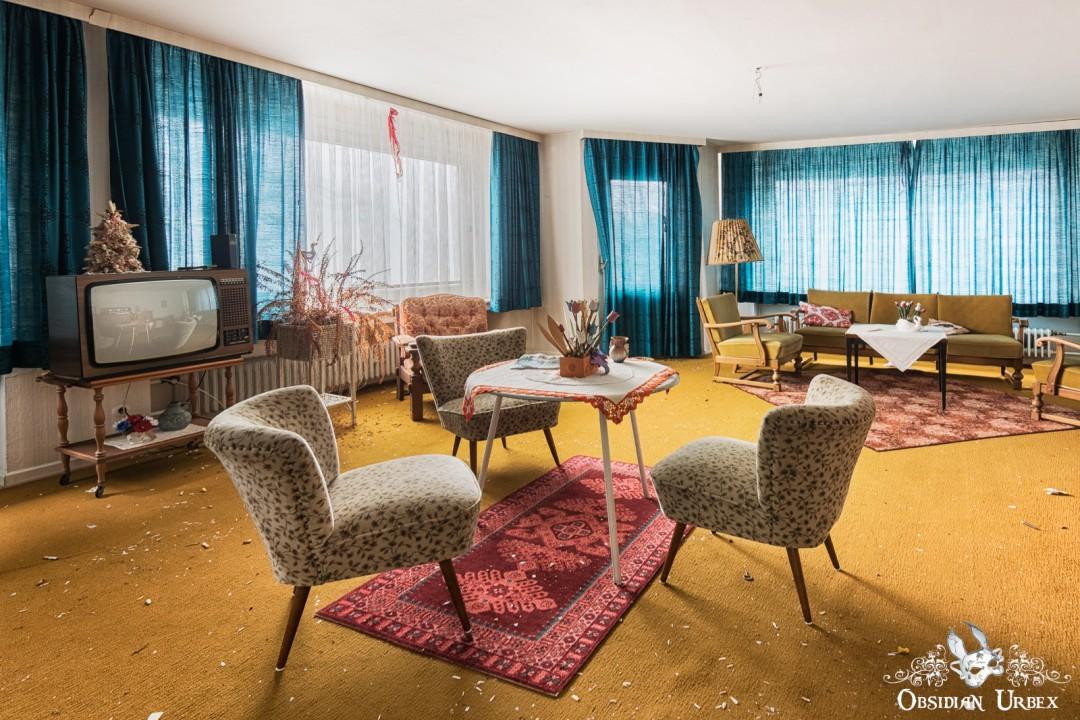 An abandoned Hotel Kurpark Germany Recreation Lounge Day Room, featuring vintage 1980s furniture, a television, and a coffee table on a yellow carpet with blue curtains.
