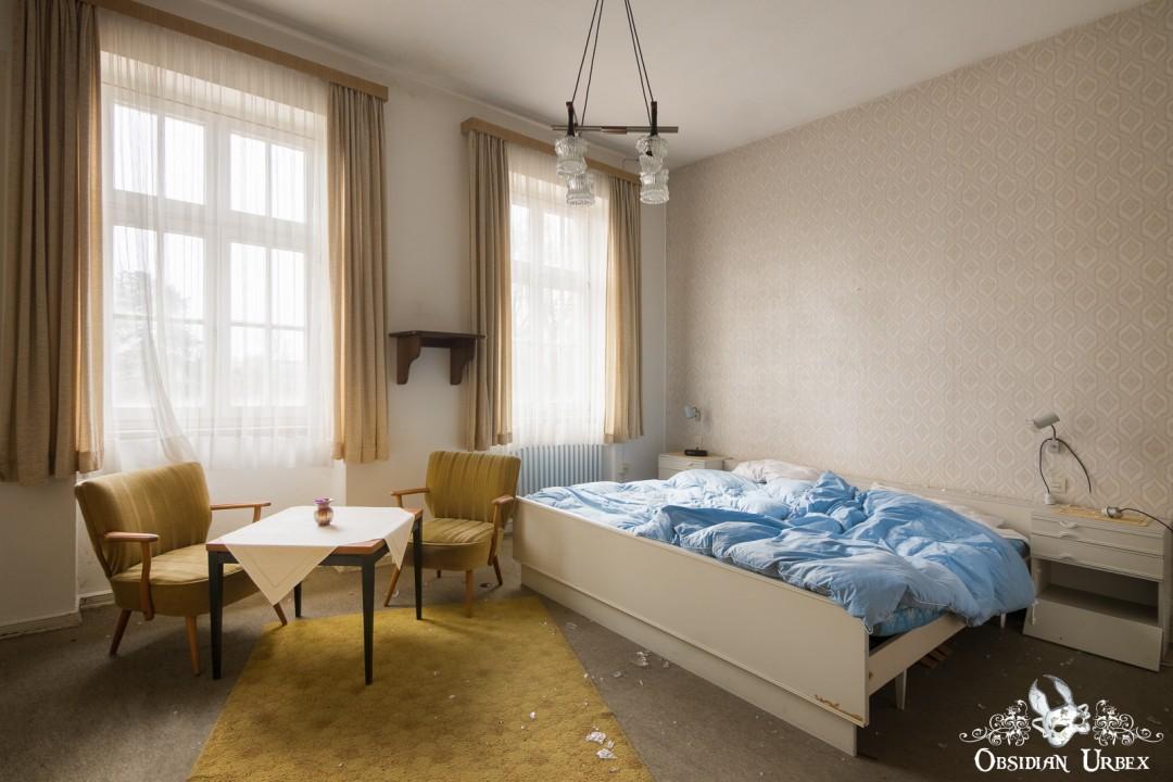 An abandoned bright hotel room from Hotel Kurpark, Germany, featuring a bed with a blue duvet, two armchairs, and large windows