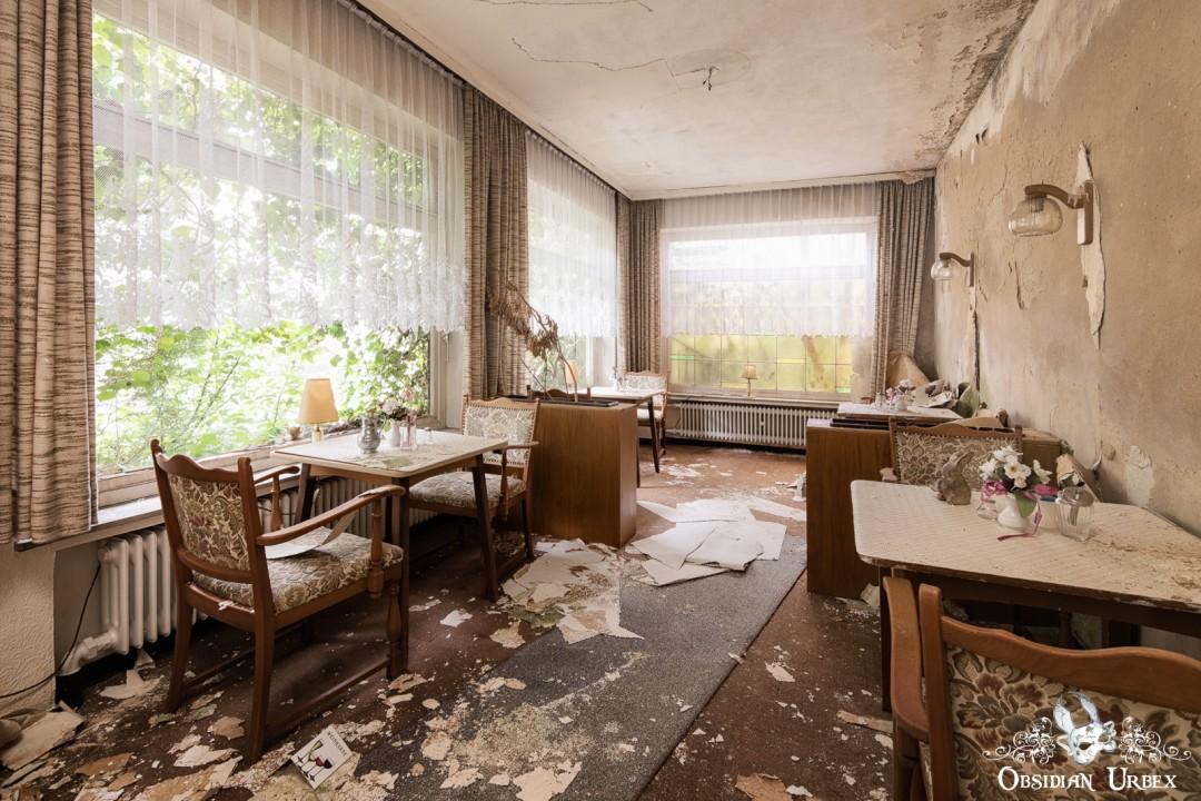 The decaying bar and breakfast area of Hotel Kurpark Germany. Shows peeling walls, debris, and old furniture with large windows overlooking greenery.
