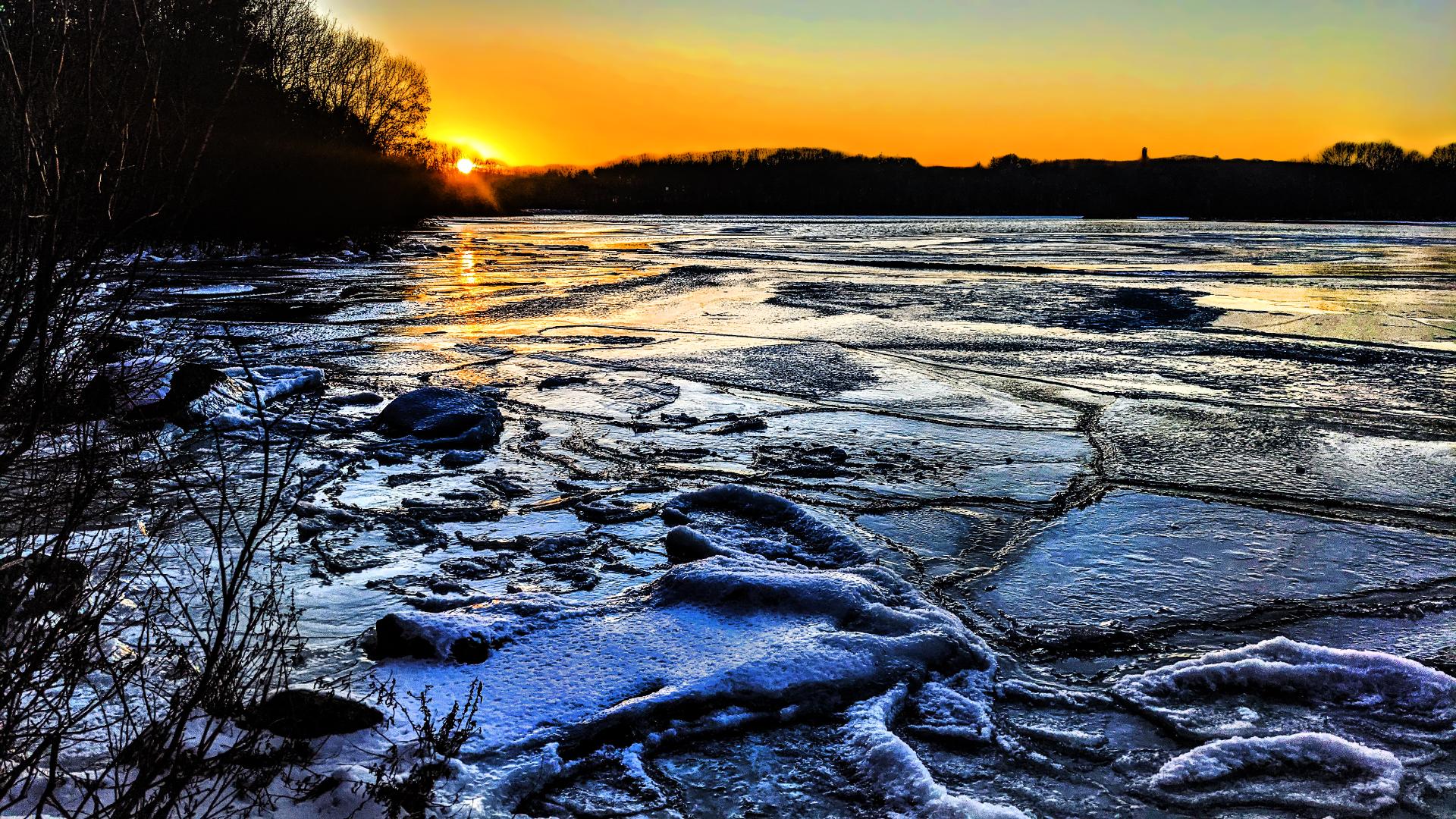 We are standing on the shore of a large frozen lake. A leafless  shrub frames the pic on the left. The visible shoreline to the left and across the lake is forest, bare leafless trees. The lake surface is frozen solid with several inches of ice thickness. There are numerous large cracks where the ice melted into pieces during thaws and then refroze back as one solid ice sheet. The sun is setting behind the trees in the far shore, tinting the sky around it and along the horizon gold. The rest of the sky is a very pale blue.