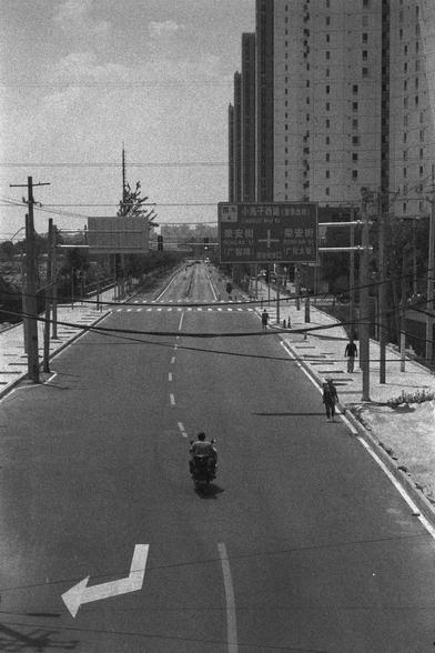Ilford HP5 Plus 400 (FF)

English
A black and white photograph of an empty street. A person on a motorcycle is visible in the middle of the road. Some people can been seen walking on the side of the street. Several high-rises can been seen in the background along some power lines. The sky is bright with some pieces of clouds.

中文
一張黑白照片. 空蕩的街道,一個人騎著一輛電動摩托車.一些人在街旁走.背景中有一些高層建築以及一些電線.天氣晴朗,有一些雲.