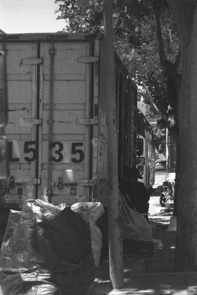 Ilford HP5 Plus 400 (FF)

English
A black and white photograph of a loading van on the side of the sidewalk. Someone on the side of the van seems to be working loading/unloading the van. Some fabrics sits in front of the van facing the camera. There is dense foliage above the van.

中文
一張黑白照片.一輛輕型貨車在人行道邊.一個人似乎在車旁裝貨/卸貨.車前有一些布料.車的上部有很厚的樹葉.