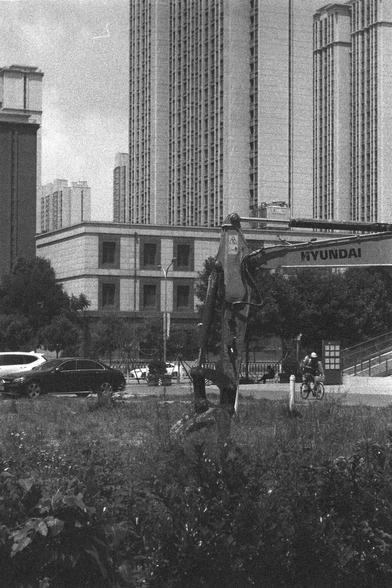 Ilford HP5 Plus 400 (FF)

English
A black and white photograph of a filed of grass with a mini-excavator. The excavator has its bucket resting in the grass. In the background stand many buildings and high-rises. A person seems to be cycling on a path along the grass field.

中文
一張黑白照片.照片中一台小型的挖掘機.挖掘機的機頭閒置在草叢當中.背景中有樓房.一個人在草叢邊的路上騎行.