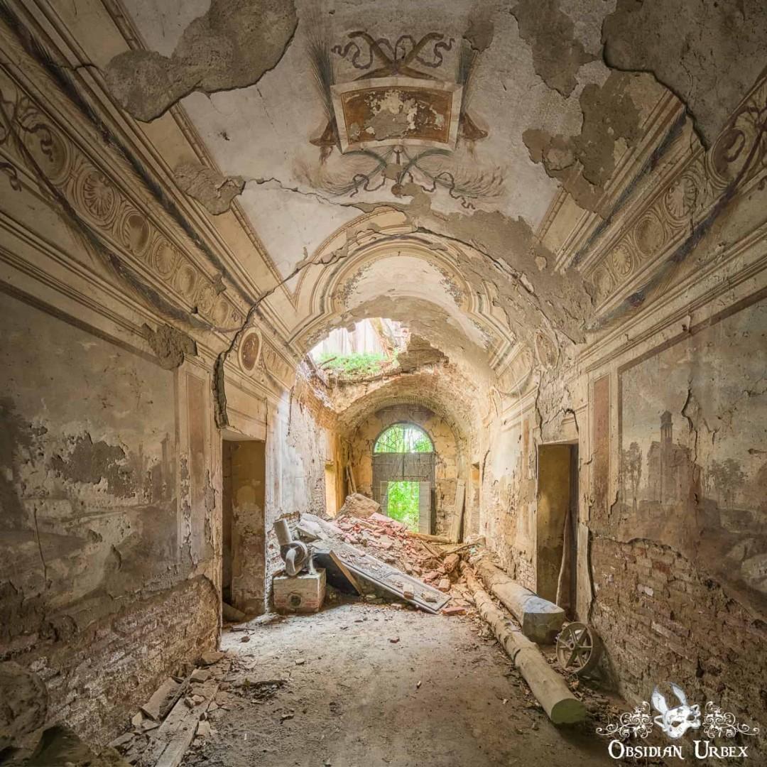 A grand, decaying hallway in an abandoned Italian villa features crumbling walls, ornate ceiling frescoes, and rubble on the floor. Light enters through an arched doorway.