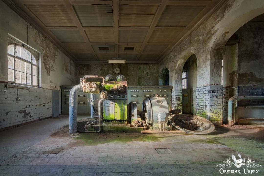 An abandoned turbine hall in a 1930s German textile factory. Decaying machinery covered in green algae sits on a tiled floor.