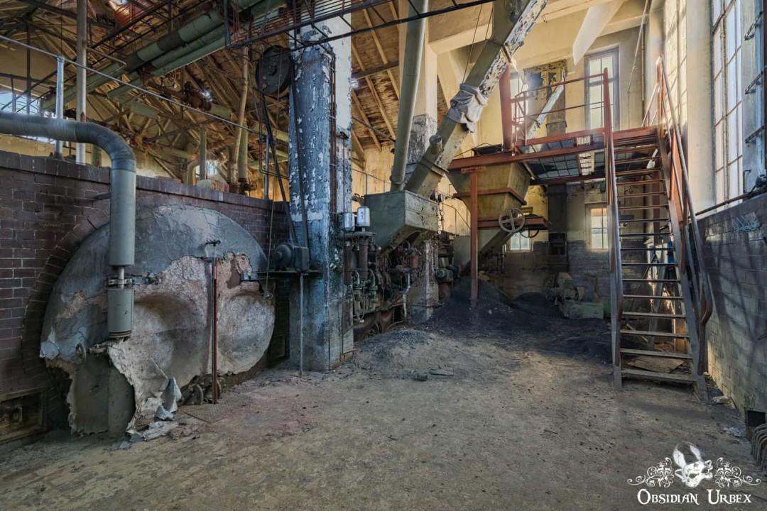 An abandoned industrial interior with large, decaying machinery, pipes, and a dusty floor.